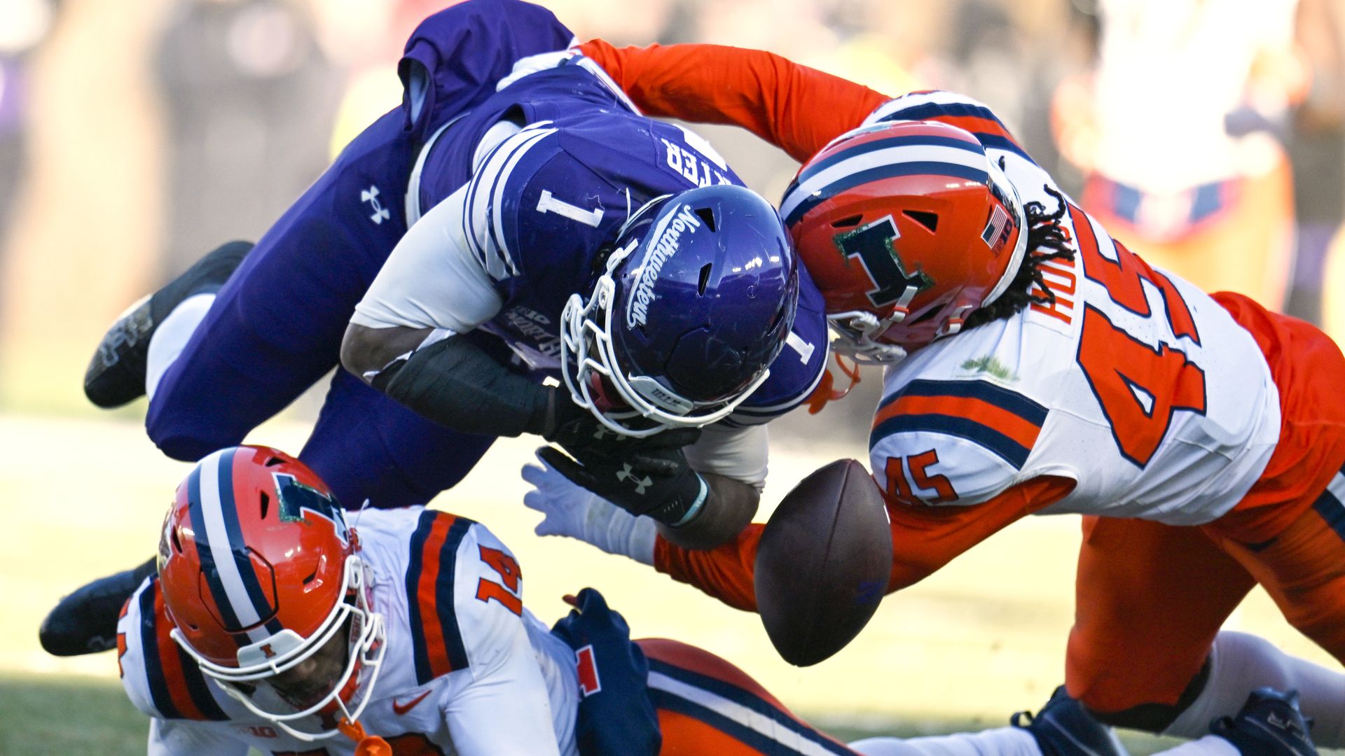 Football player in purple tackled mid-air by two players in white jerseys with orange helmets, one on ground and one lunging, football flying between them on grassy field.