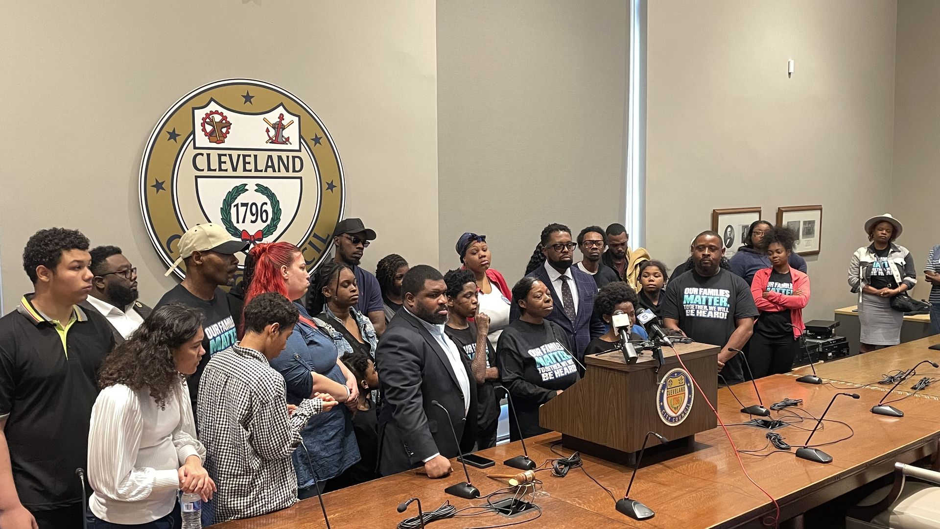 A diverse crowd stands in a Cleveland City Council chamber beside a large Cleveland seal on the wall; a podium with microphones sits at the table, attendees wearing teal shirts.