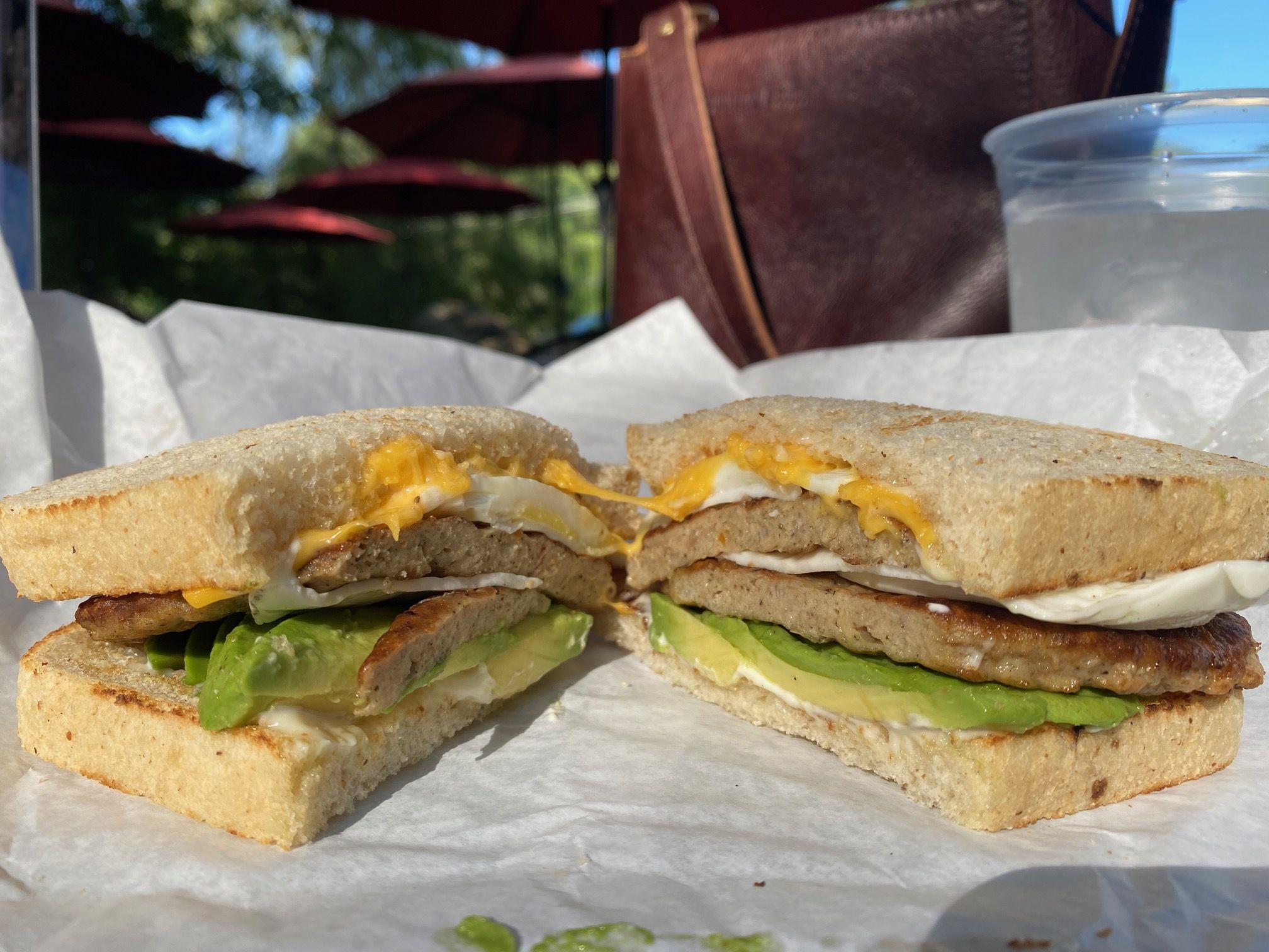 Sandwich cut in half with sausage patty, slice of avocado, fried egg with melted cheddar cheese, and mayo between two pieces of toasted white sandwich bread on white paper.
