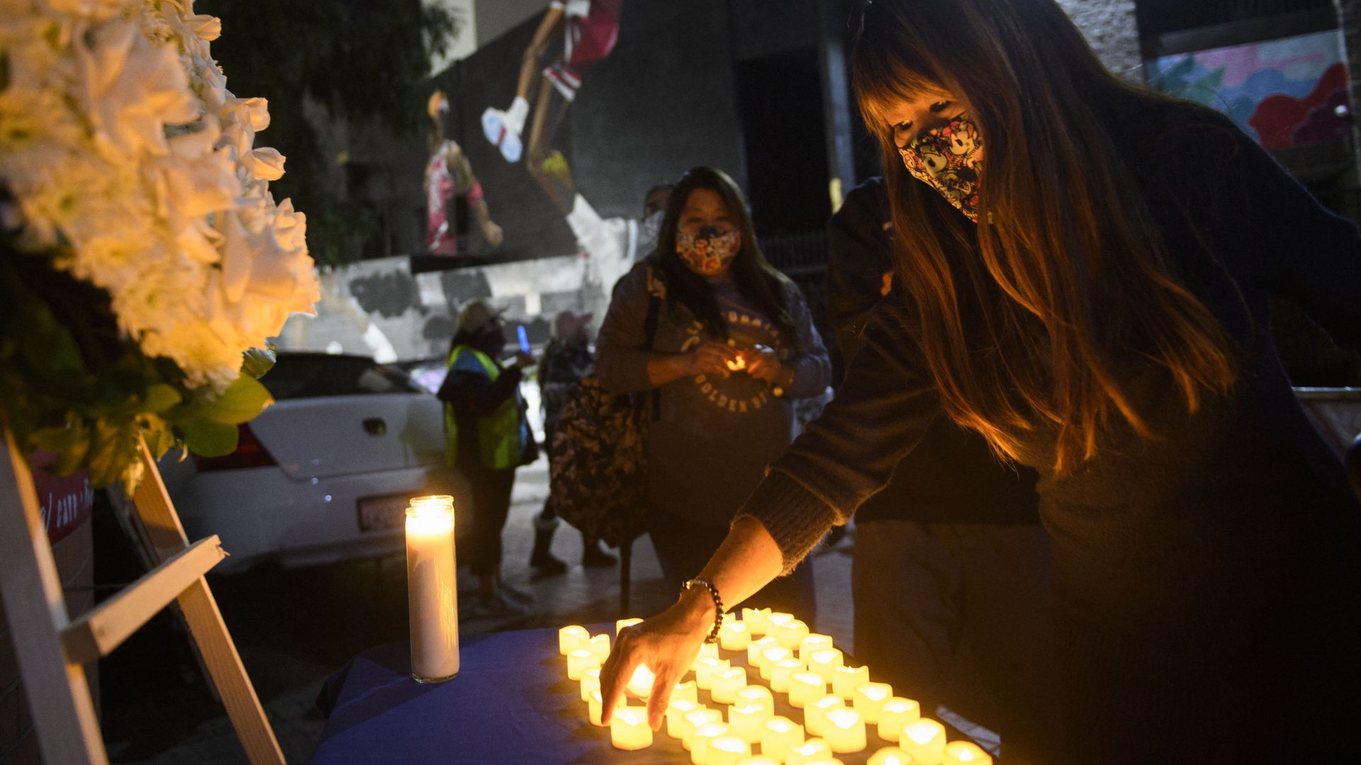 Photo of a masked person placing an electric candle in front of a white flower shrine at a COVID vigil