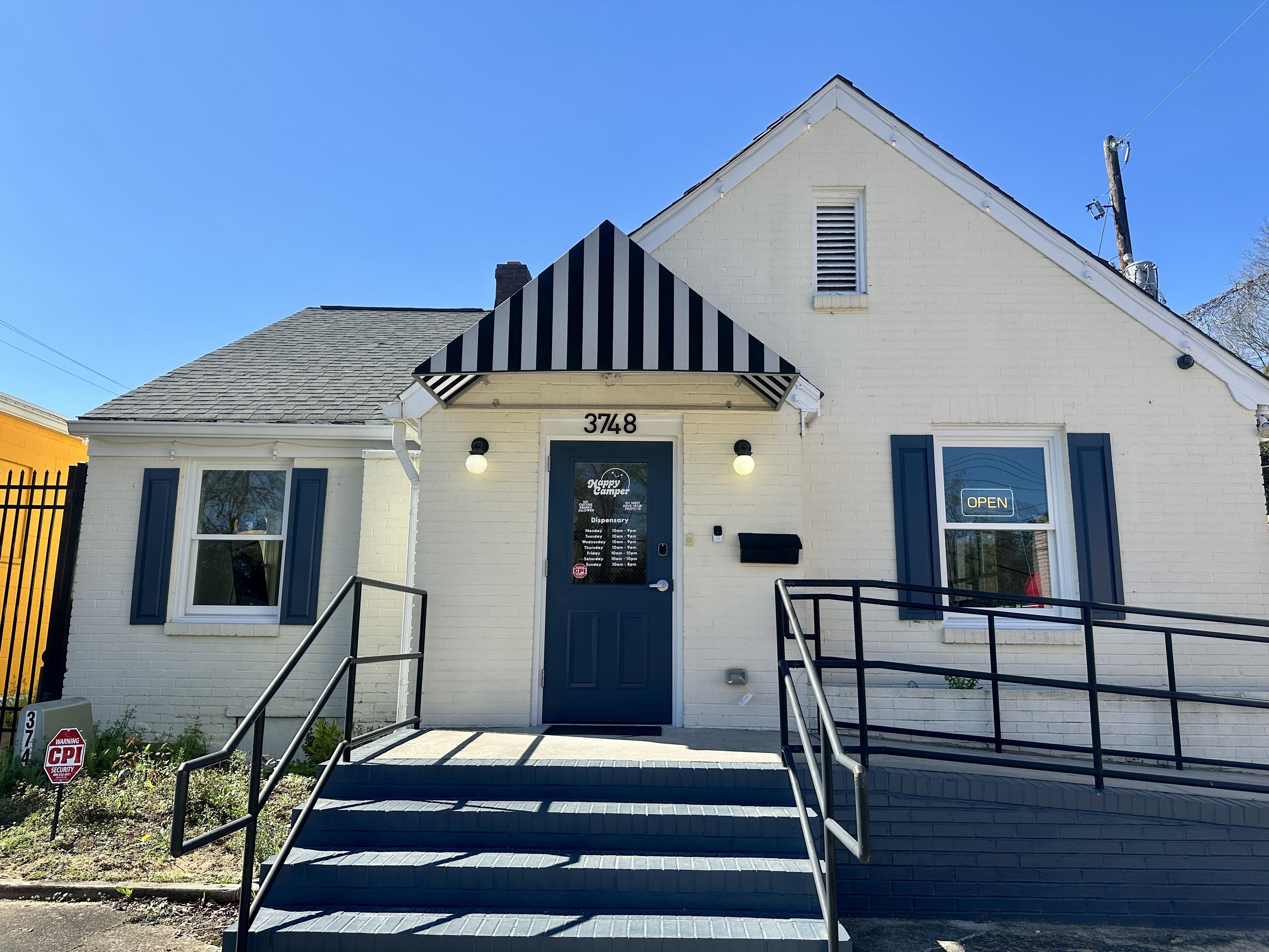 White brick building with a blue door under a black-and-white striped awning, address 3748. Two porch lights flank the door, blue shutters, an "OPEN" sign in the right window, and ramps.