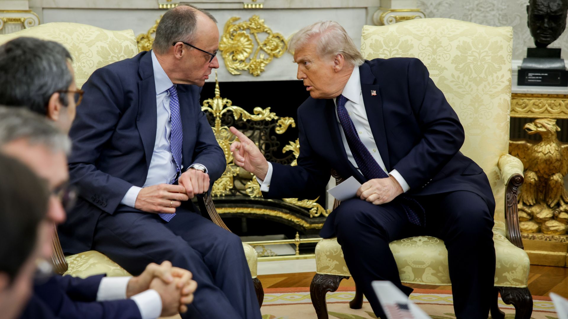 Two men in dark suits lean close, speaking in a luxurious cream-and-gold room with ornate chairs and a marble fireplace; a bust is visible in the background as one gestures with a raised finger.