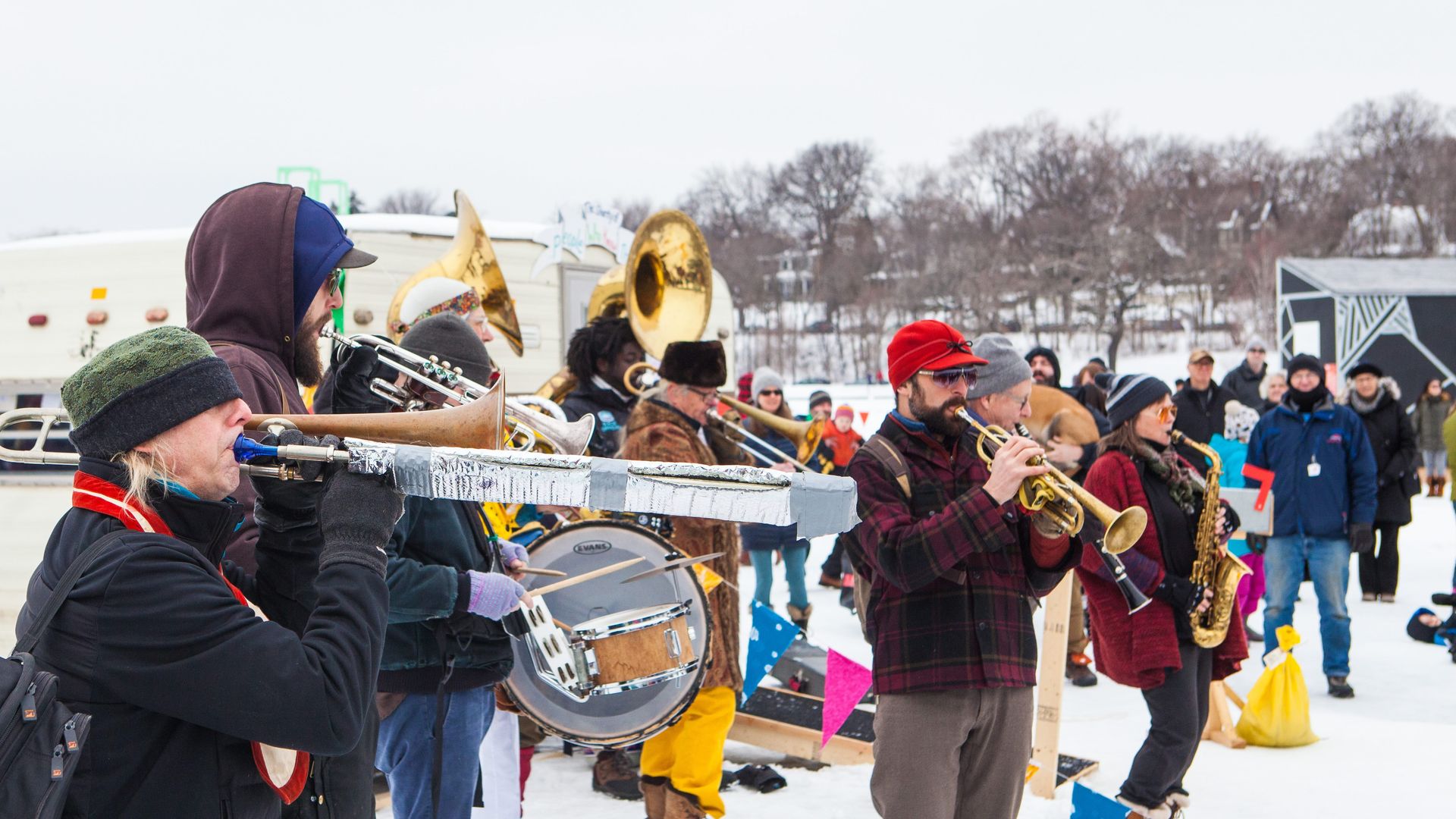 Outdoor winter scene with a diverse group of musicians playing brass and woodwind instruments while bundled in warm clothing. Snow covers the ground, and a crowd watches in the background.