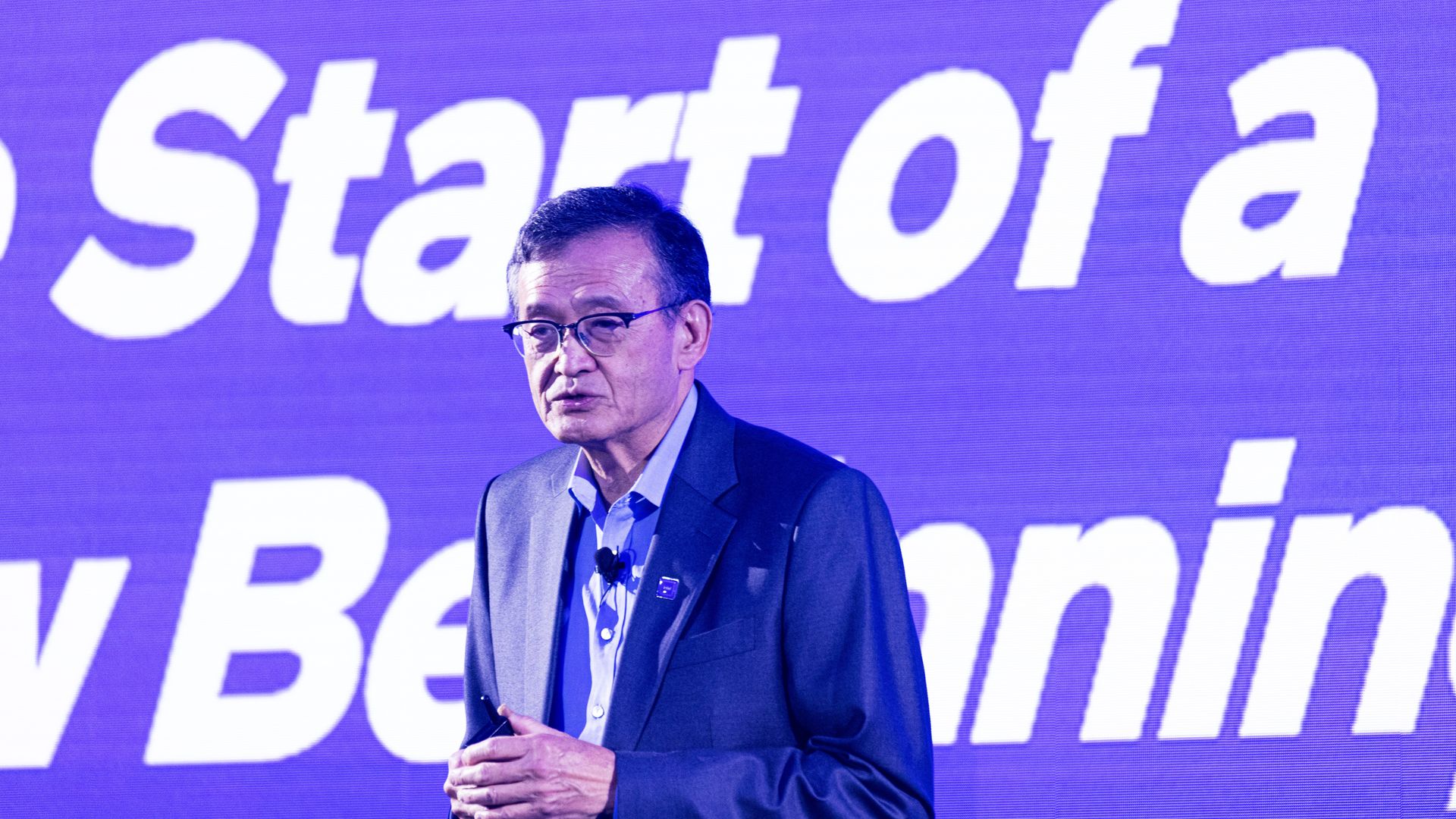 A person in a gray suit and glasses speaks in front of a purple screen displaying large white text starting with "Start of a".