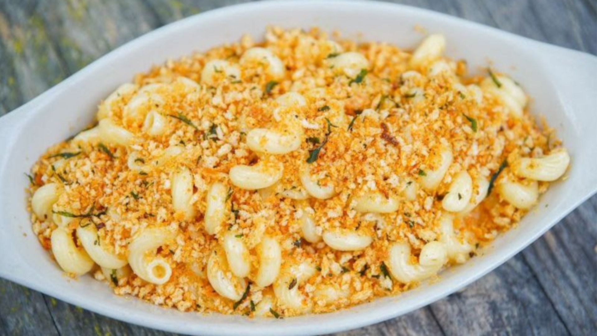 White oval baking dish of baked pasta shells topped with golden breadcrumbs and cheese, sprinkled with herbs on a rustic wooden surface.
