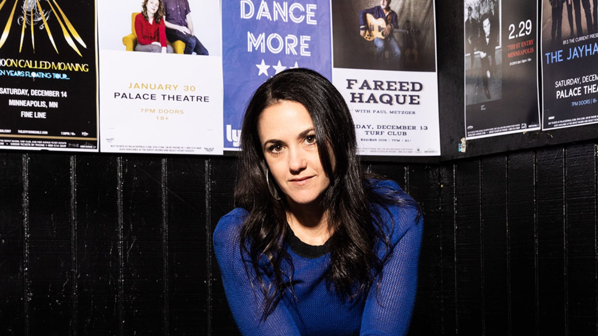 A photo of Dayna Frank in front of a wall covered in concert posters.