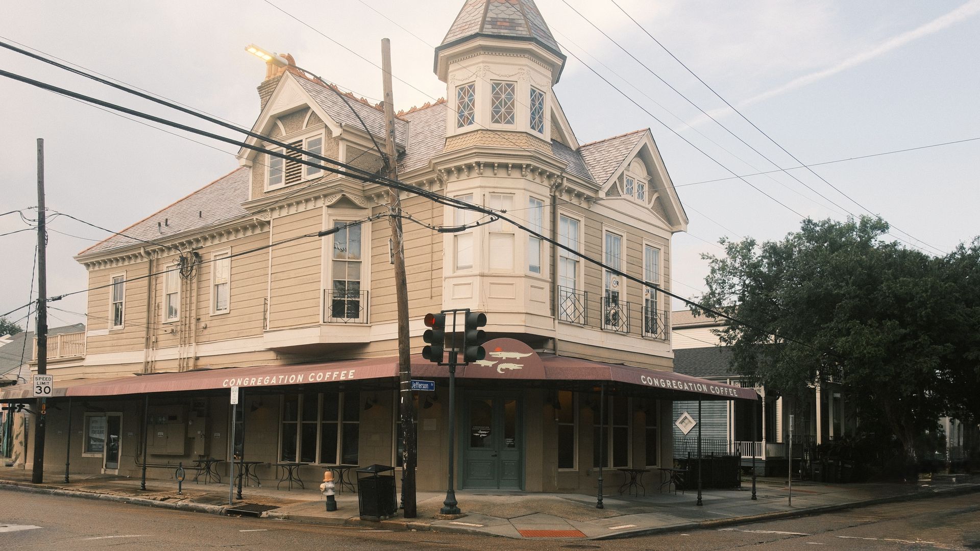 Two-story beige Victorian building with a turret houses Congregation Coffee cafe. Red awning, green double doors, outdoor tables, traffic light, and street signs visible at wet intersection.