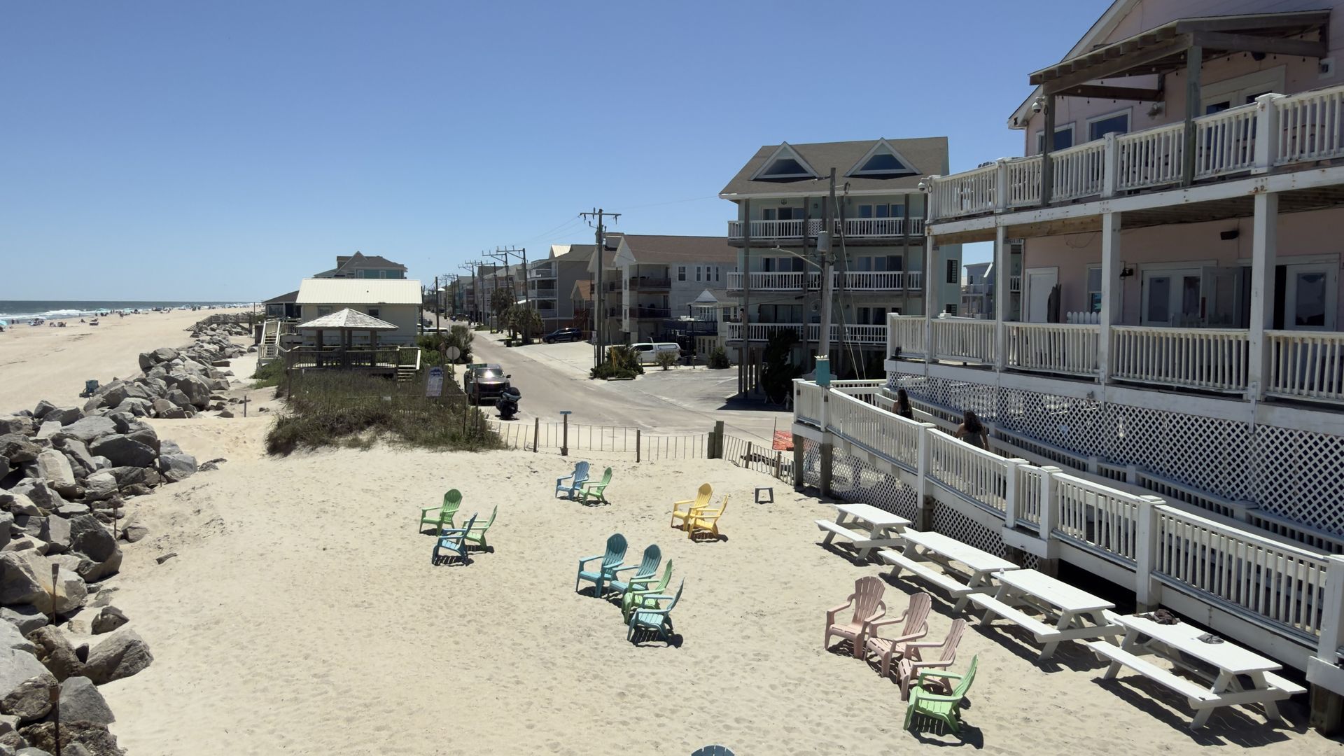 Bright beachside scene with a clear blue sky and a white contrail. Left: sandy beach with a rocky jetty; center: colorful chairs; right: stacked beach houses with white railings along a street.