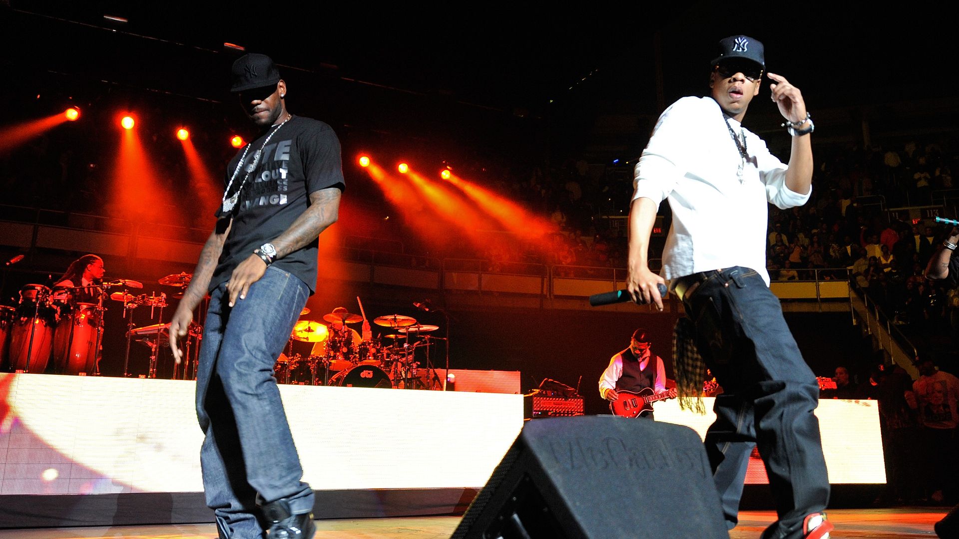 Two male performers on a stage at night under orange lights; both wear NY caps, one in a black T-shirt and jeans, the other in a white shirt. Drums and guitarist visible, audience in the stands.