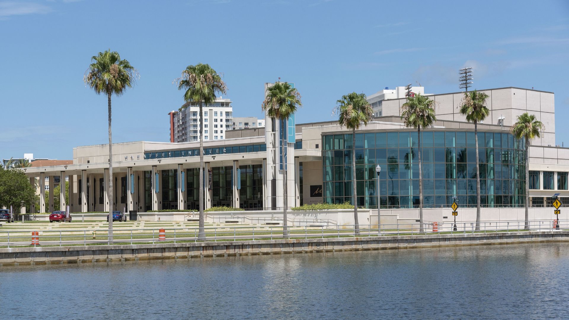 A modern riverside campus building with a curved glass facade and beige walls, flanked by tall palm trees. A calm waterway runs along the foreground under a clear blue sky.