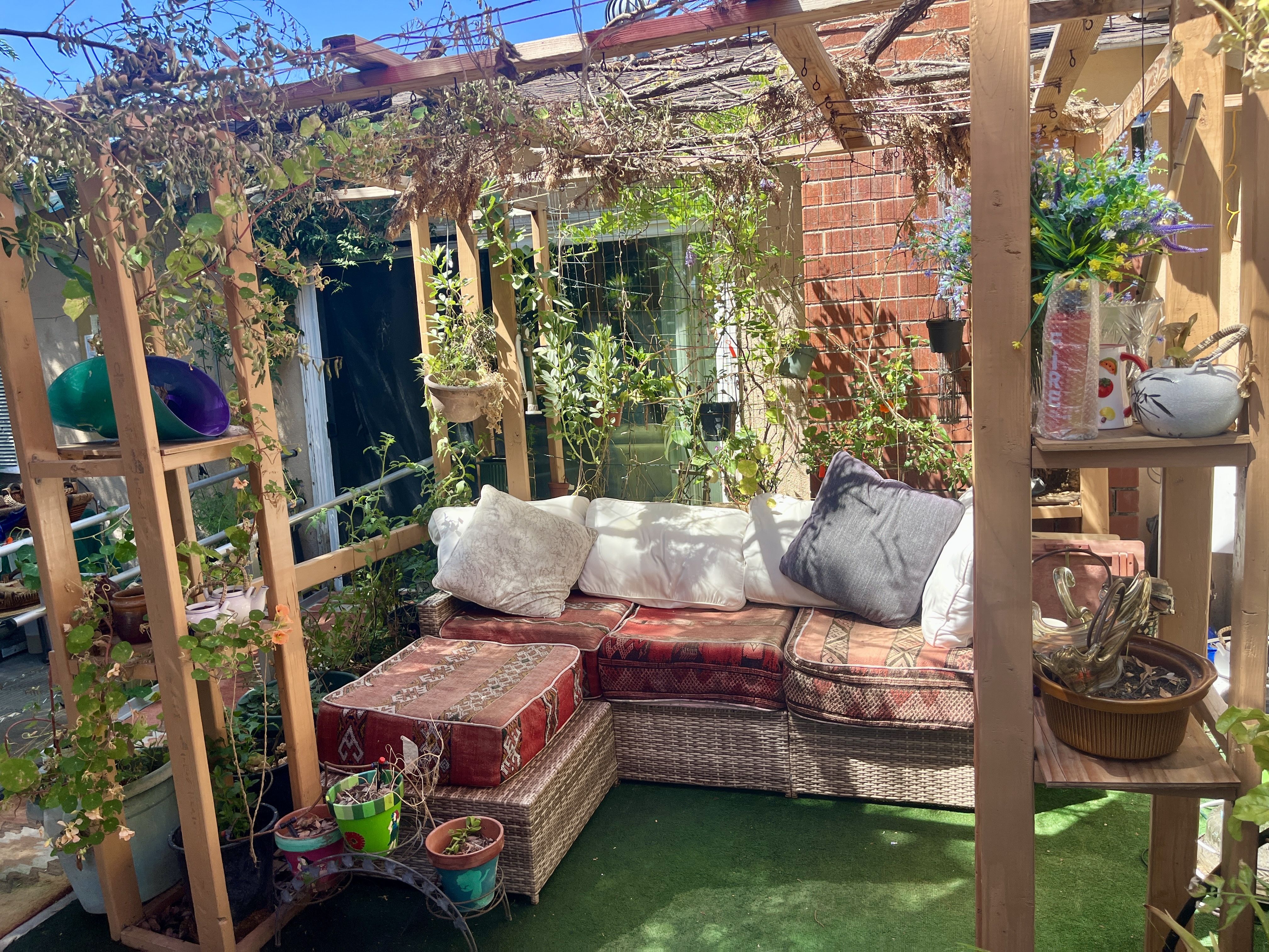 Cozy outdoor seating under a wooden pergola with climbing vines, a wicker sofa with red patterned cushions, potted plants, and a brick wall adorned with flowers.