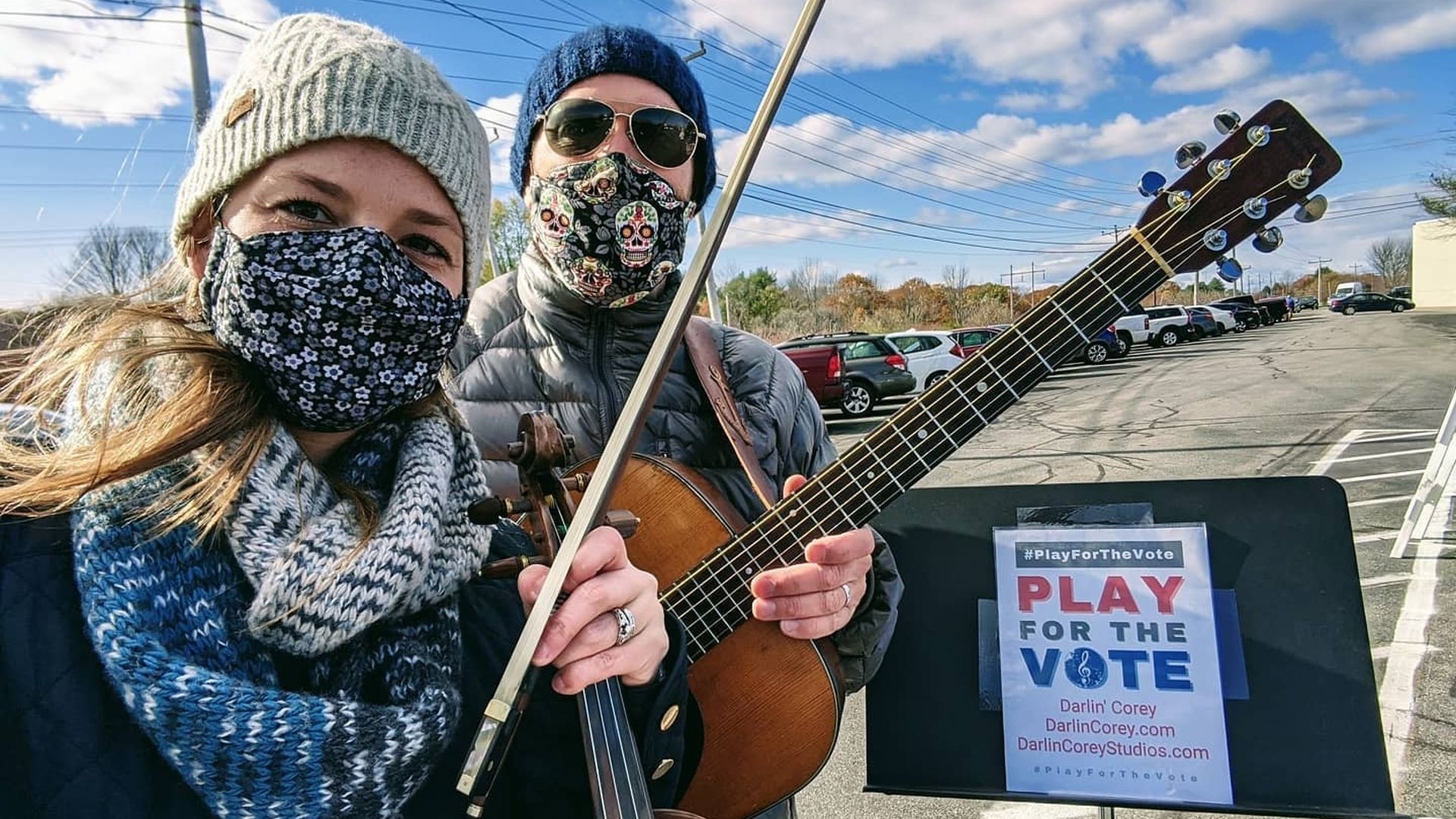 A woman wearing a face mask holding a violin and bow next to a man in a face mask holding a guitar next to a music stand with a paper that reads "Play the Vote."
