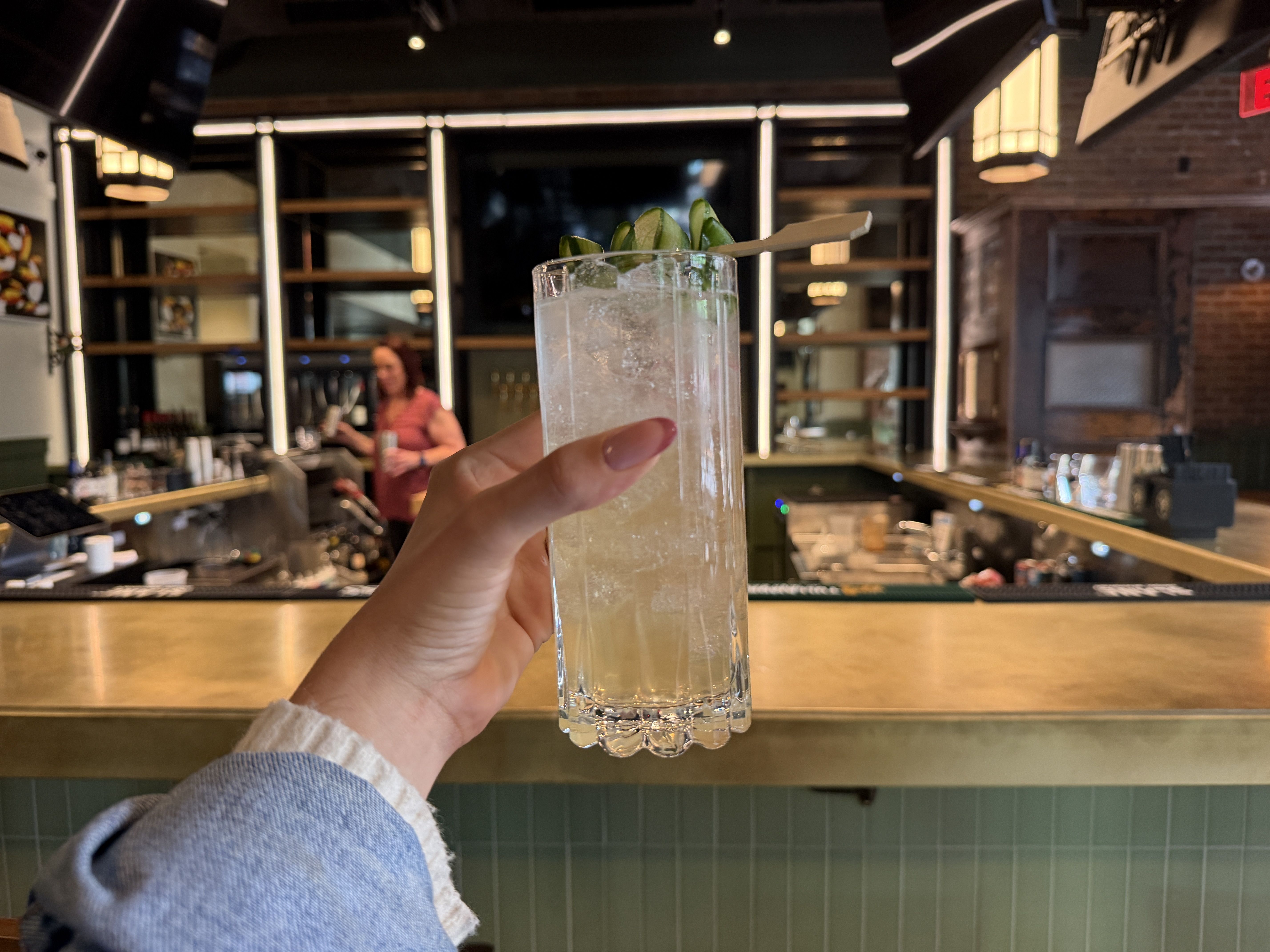 A hand with pink nails grips a tall, clear glass of iced cocktail topped with lime wedges, in a warmly lit bar with a bartender and shelves blurred in the background.