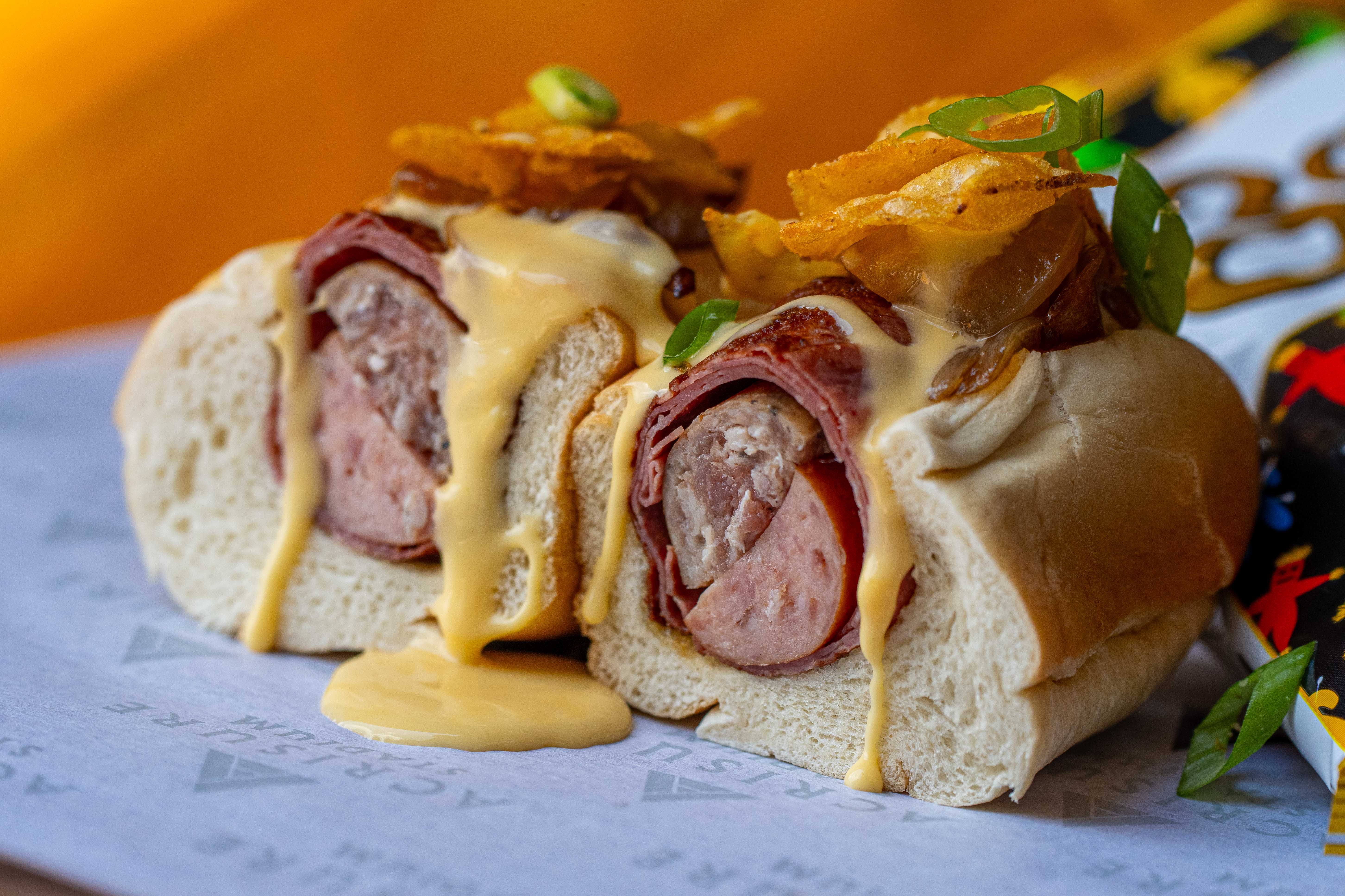 Close-up of a italian sausage and cheddar bratwurst on a soft roll, layered capicola, melted cheese sauce, crispy potato chips, with extra sauce dripping onto the plate.