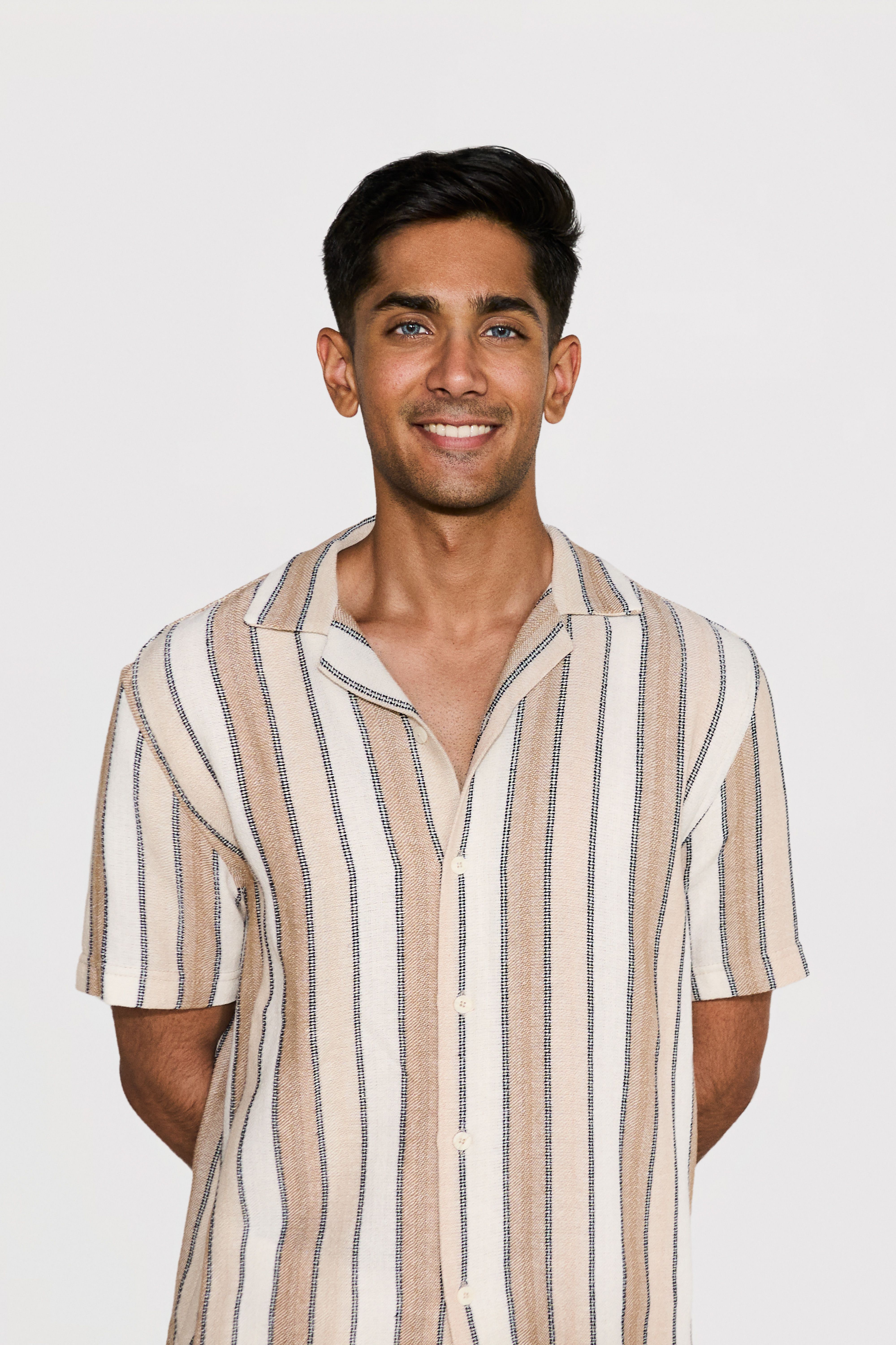 Smiling young man with dark hair and blue eyes wearing a beige, white, and black vertical striped short-sleeve button-up shirt, standing against a plain white background.