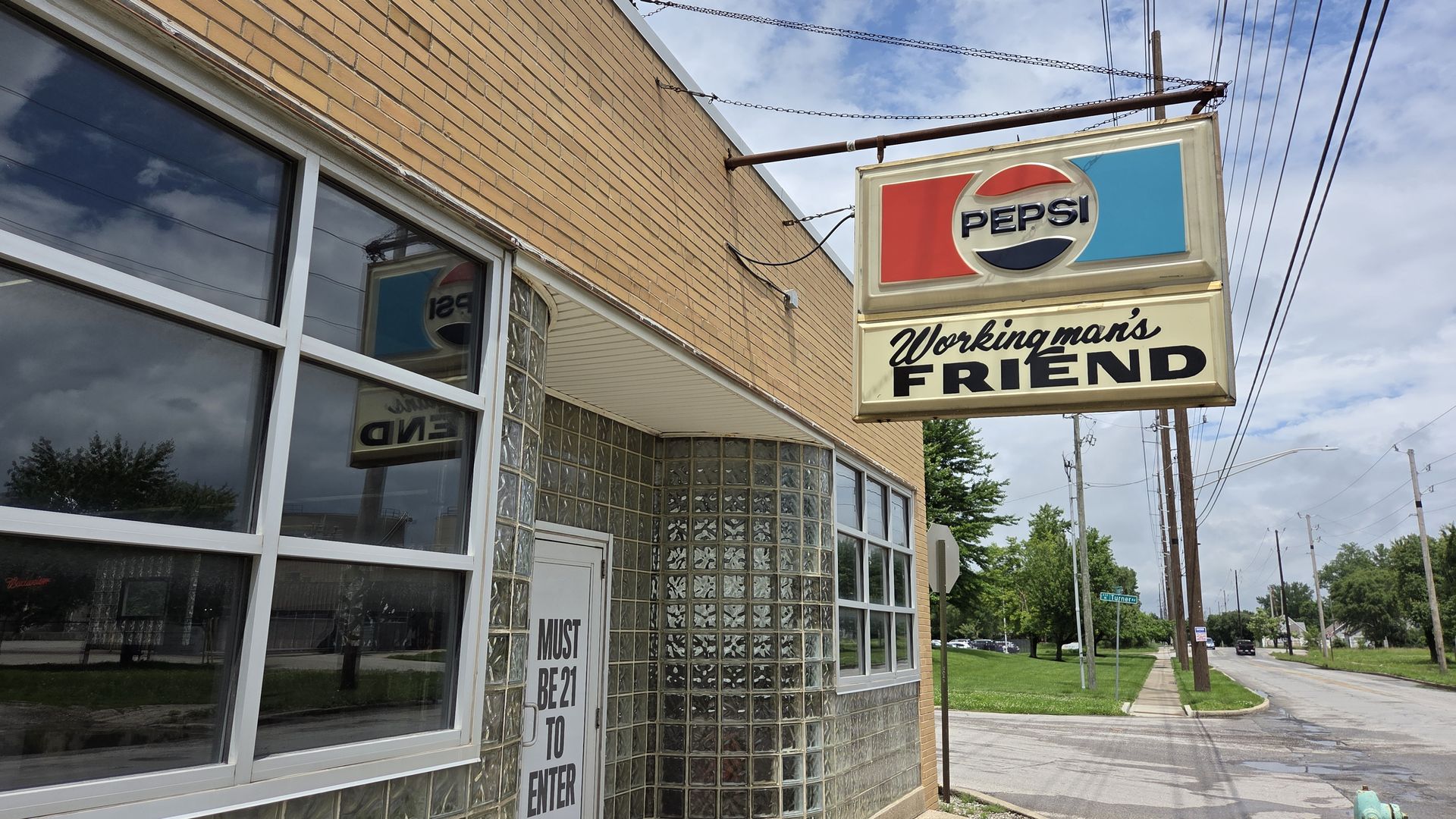 Outdoor view of a brick storefront with a hanging Pepsi sign in red, white, and blue; the sign reads "PEPSI" and "Workingman's FRIEND" in cursive, under a blue sky with power lines.