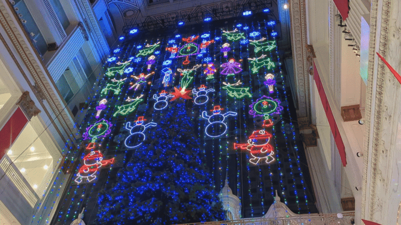 Bright neon holiday lights depicting snowmen, reindeer, wreaths, and angels in blue, red, green, and purple, hanging inside a large, ornate building with cream-colored walls and red banners.