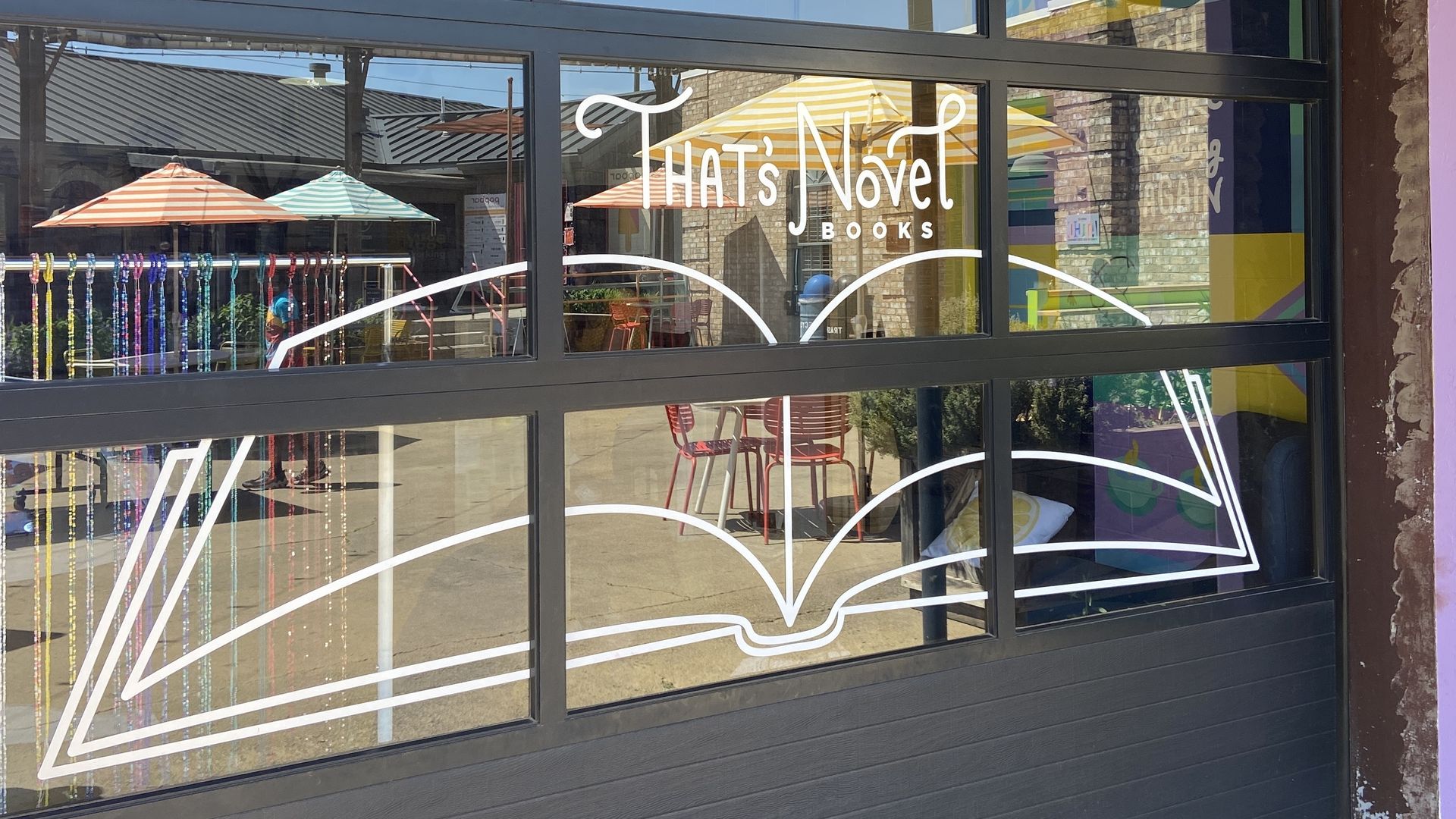 Colorful bookstore storefront with large glass windows. Pink wall above features green circles and yellow shapes; white line art of an open book and the sign "That's Novel Books" inside.