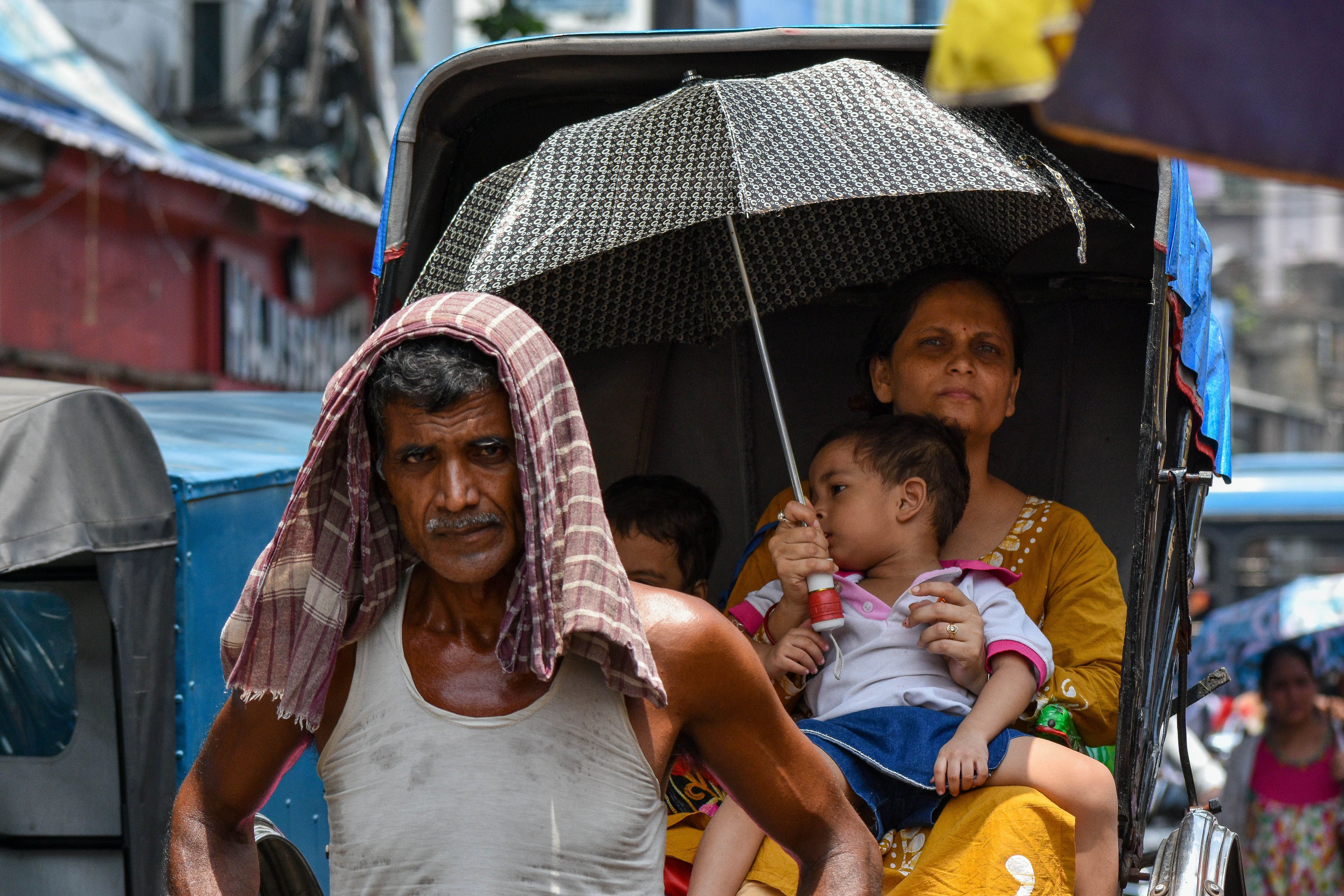 A man pulling a rickshaw wearing a cloth over his head due to the heat. The passengers in the rickshaw, a mother and her children, are using an umbrella to stay in the shade. 