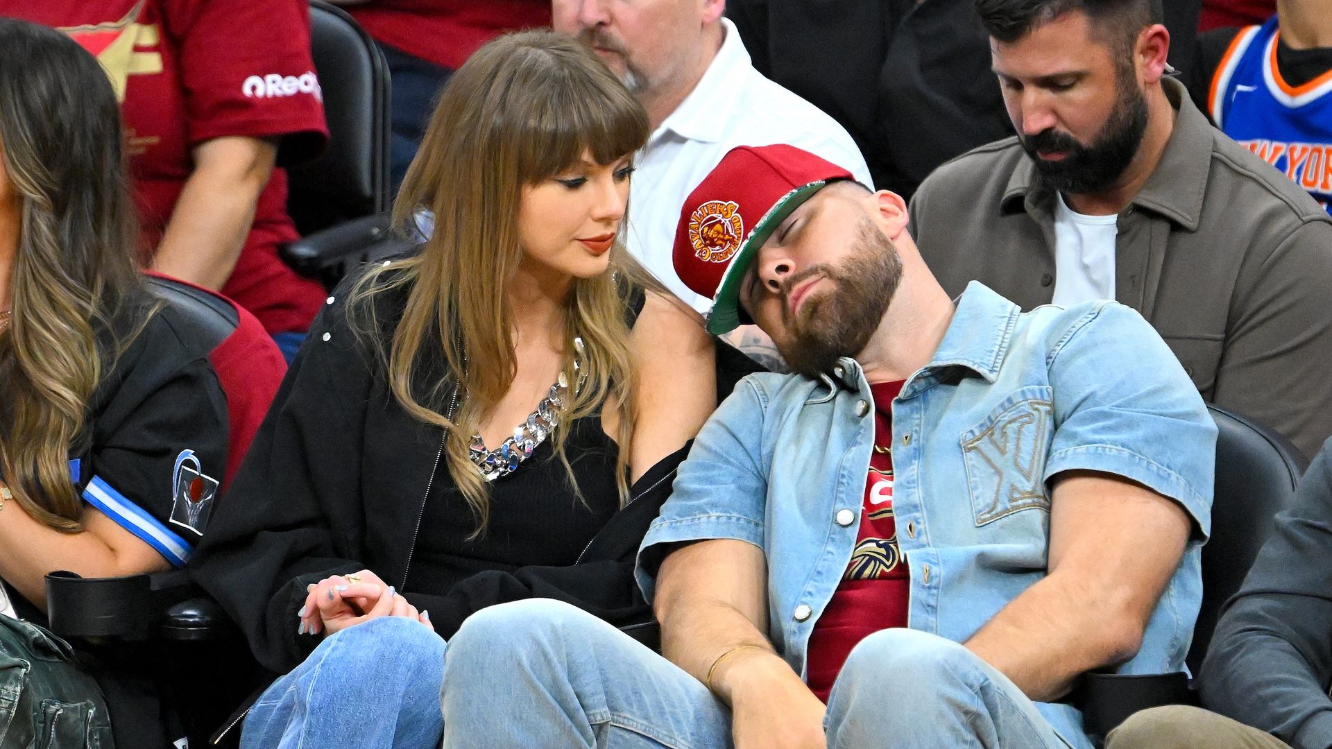 Two spectators in a crowded arena: a woman with long brown hair in a black jacket and chunky necklace gazes at a man in a denim shirt who leans on her shoulder; a bearded man sits nearby.