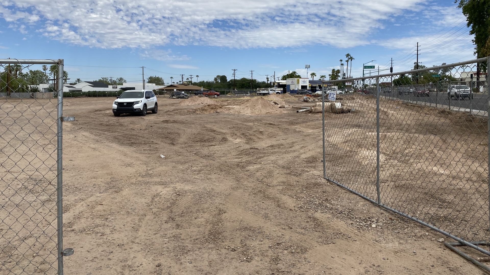 A white car on a dirt lot surrounded by a chain link fence with an intersection in the background.