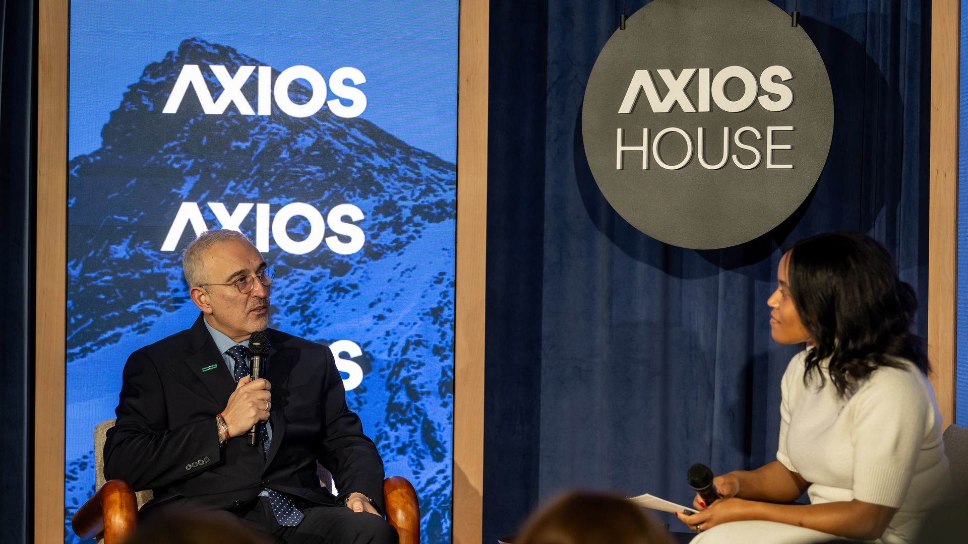 Two people having a discussion on stage at Axios House, with a man in a suit holding a microphone and a woman in a white dress listening and holding a microphone and notes.