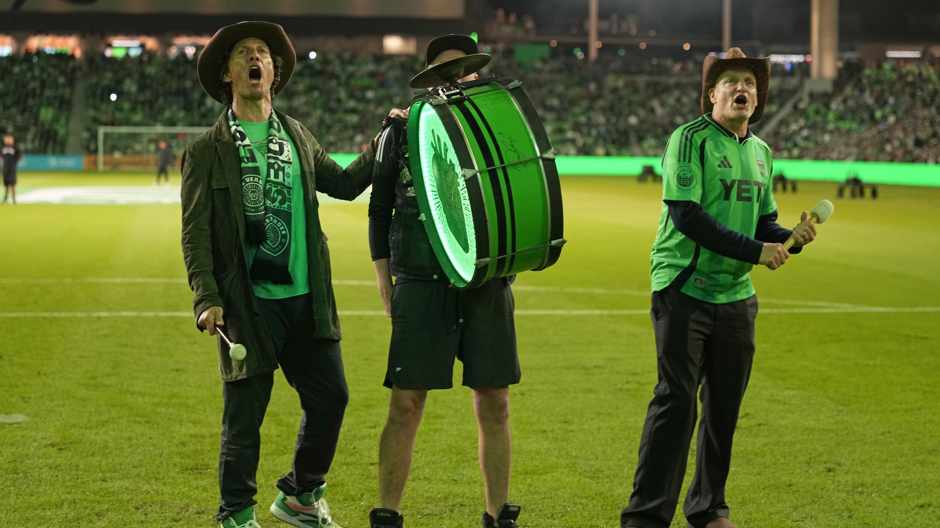 Three enthusiastic men on a soccer field at night, two wearing cowboy hats and green clothing, one playing a large green drum with a glowing LED light, cheering in front of packed stadium.