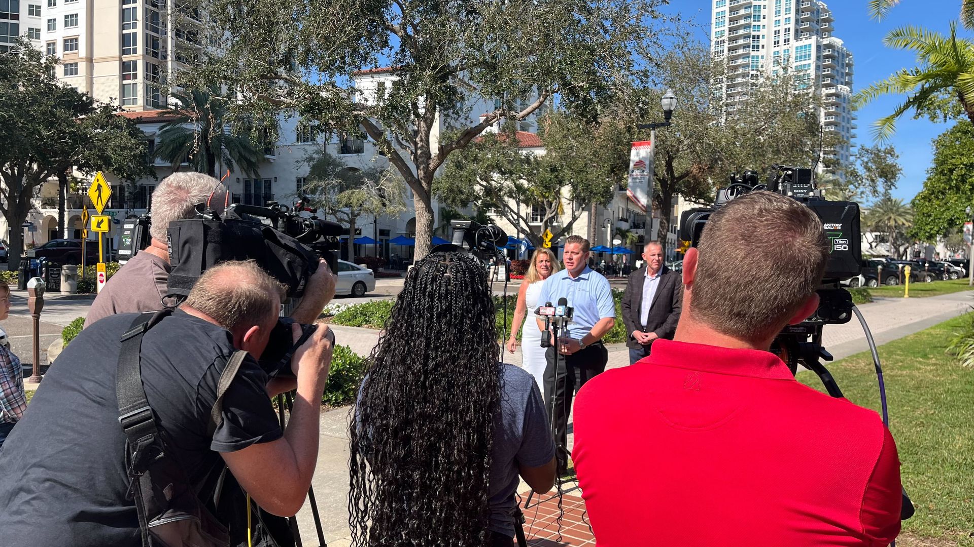 A group of journalists point their cameras toward a man in a light blue polo shirt and dark slacks standing at a microphone stand. In the background is a Mediterranean-style building flanked by high-rises.