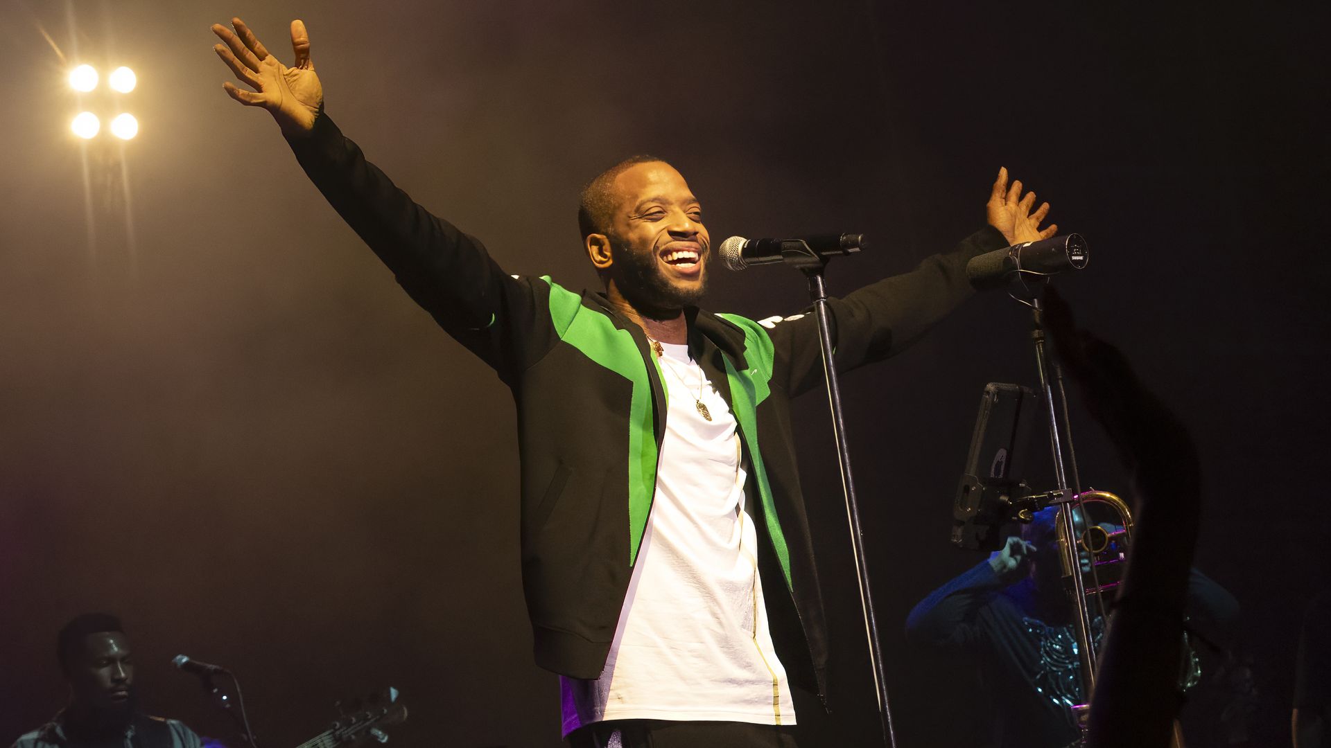 Trombone Shorty performs onstage, smiling in front of a microphone with both arms outstretched.