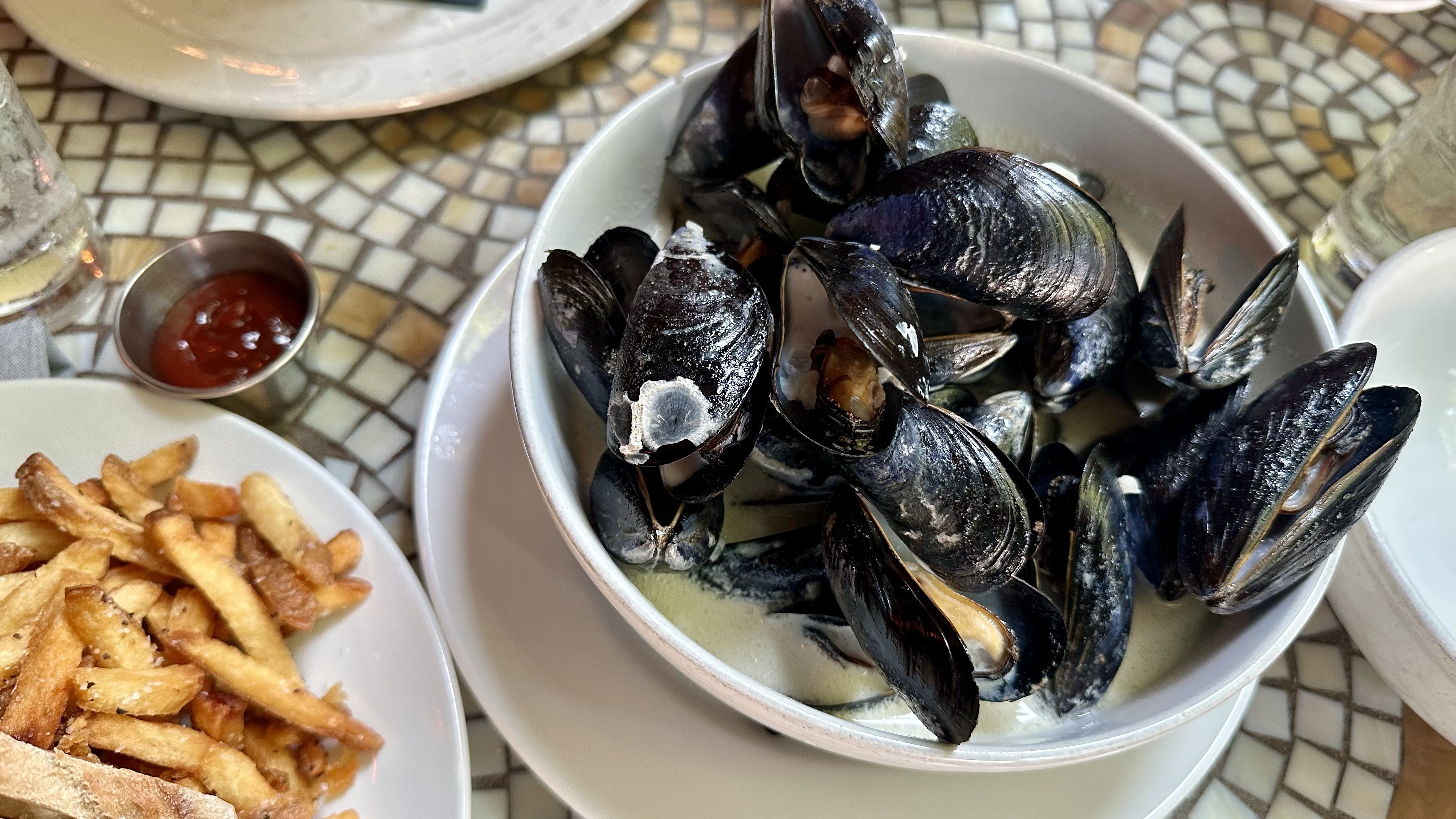 Mussels in dark shells in a white bowl with pale broth; some meat visible. French fries and a small ketchup cup sit nearby on a mosaic table.