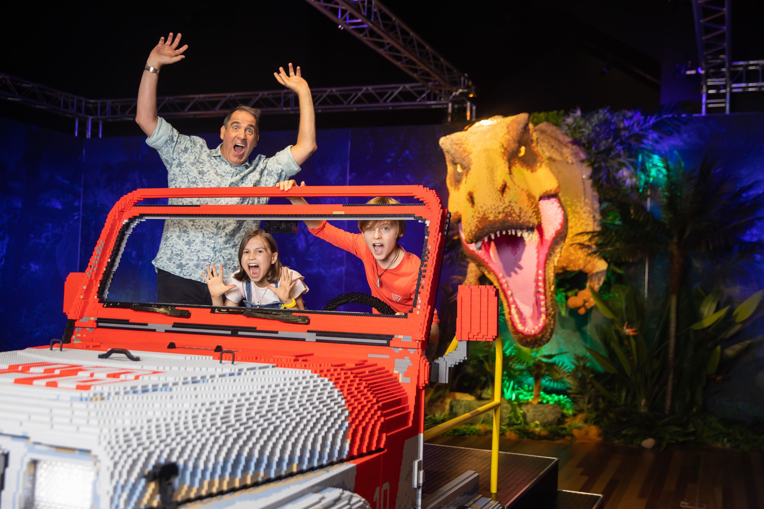 Three people ride a red LEGO-style vehicle on a stage, with waving hands; a large roaring dinosaur prop looms behind them under blue and purple lights with tropical plants.