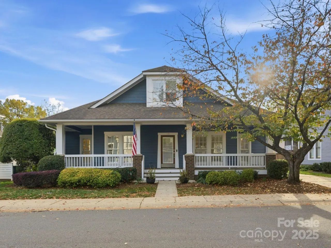 Blue single-story house with white trim, front porch, American flag, bushes, a tree with autumn leaves, and a clear blue sky background.