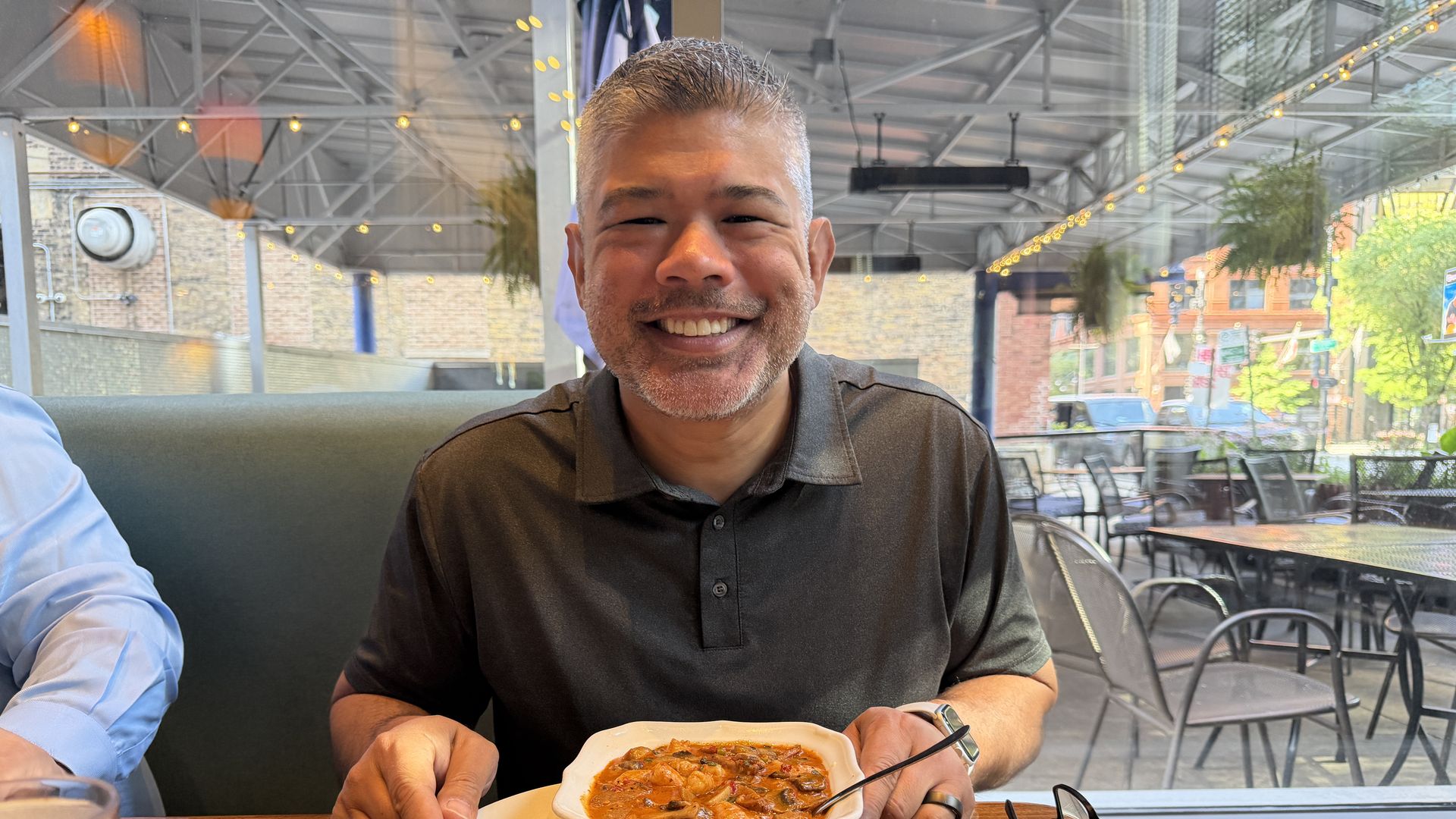 Smiling man with short gray hair and stubble sits at a wooden table in a bright cafe, with bowls of curry and a bread roll before him; string lights, glass walls, and brick buildings visible outside.