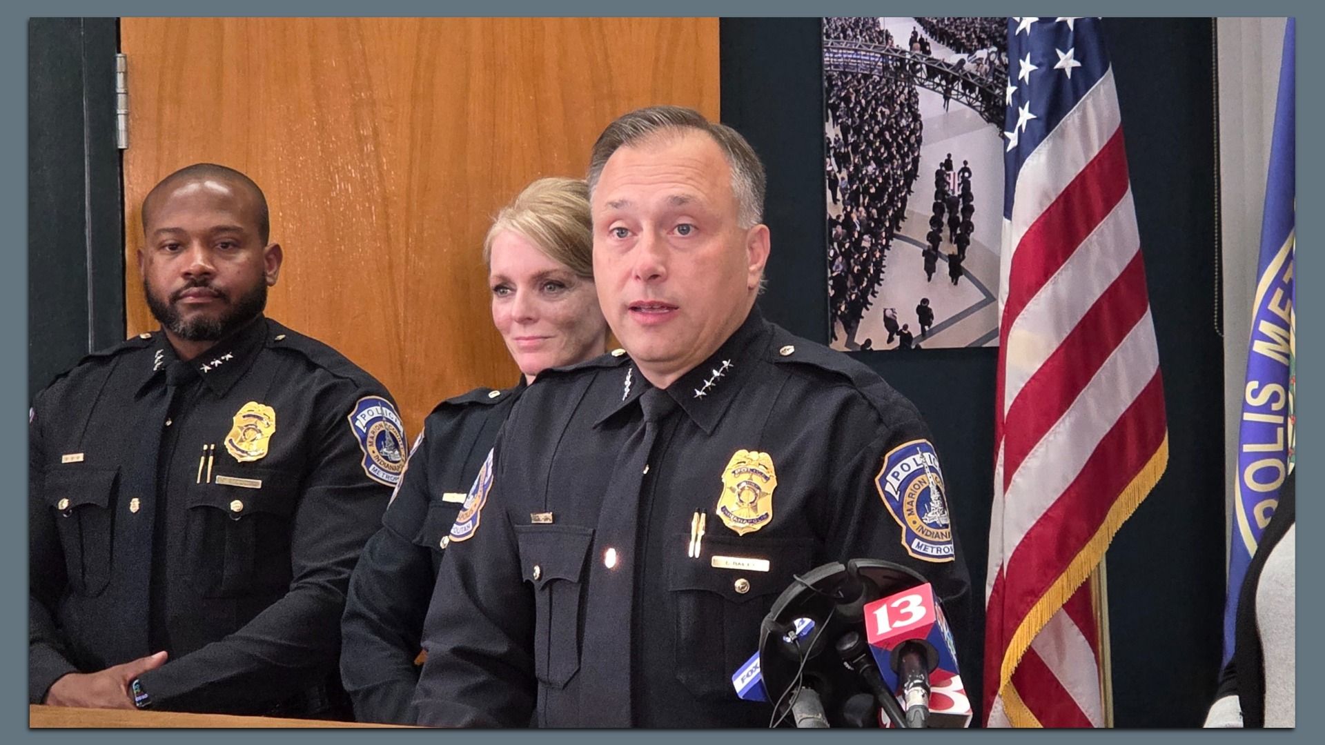 Three police officers in black uniforms at a press conference, with an American flag and police department flags in the background, and microphones in front of them.