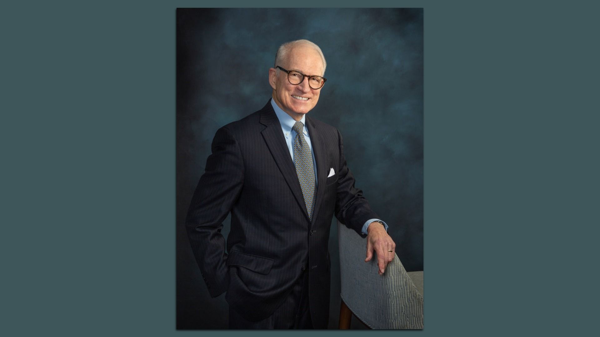 Portrait of an older man with gray hair and glasses, wearing a dark pinstripe suit, light blue shirt, patterned tie, and a white pocket square, posed beside a chair against a teal backdrop.