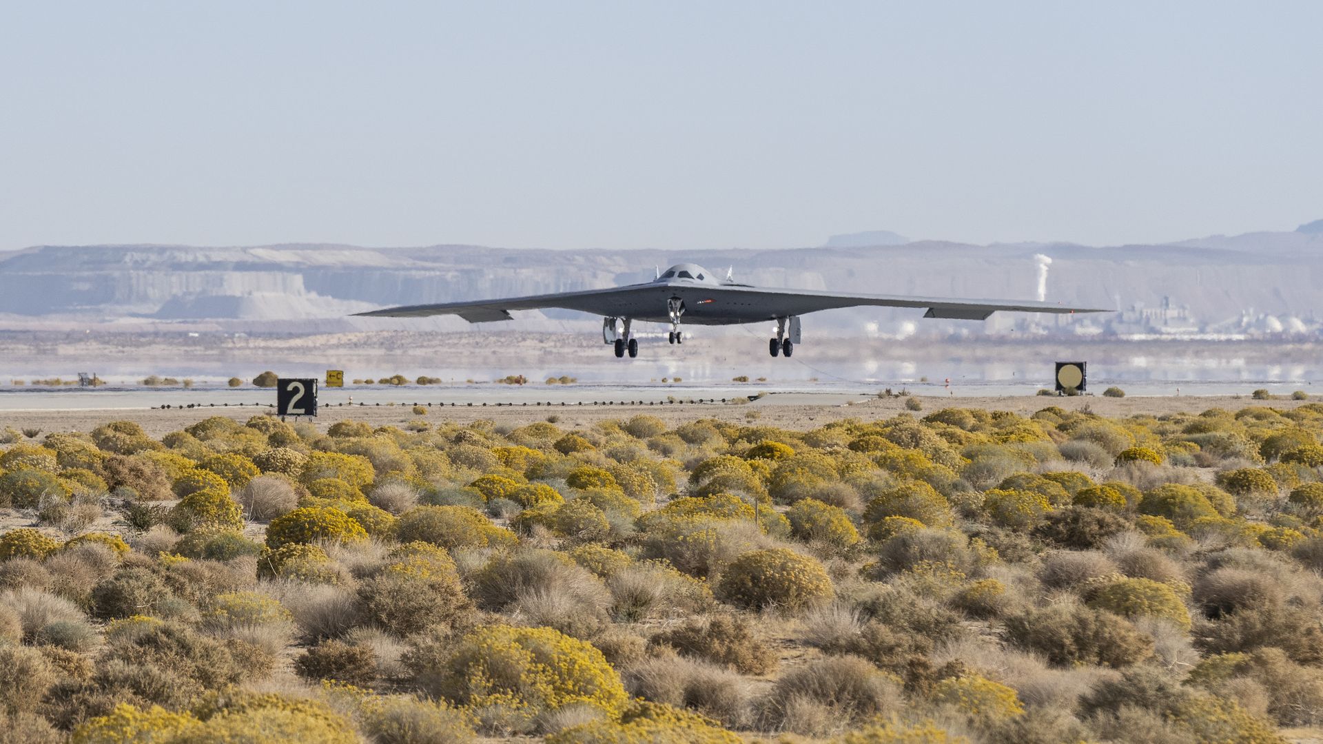 A B-21 Raider lands on a runway. The foreground is scrublands. The background is hazy with heat.