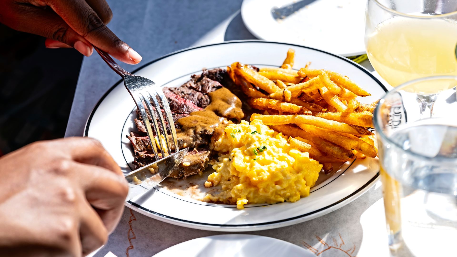 Hands holding fork and knife cutting steak on plate with brown gravy, scrambled eggs, and golden French fries, with a glass of water and a yellow drink on the side.