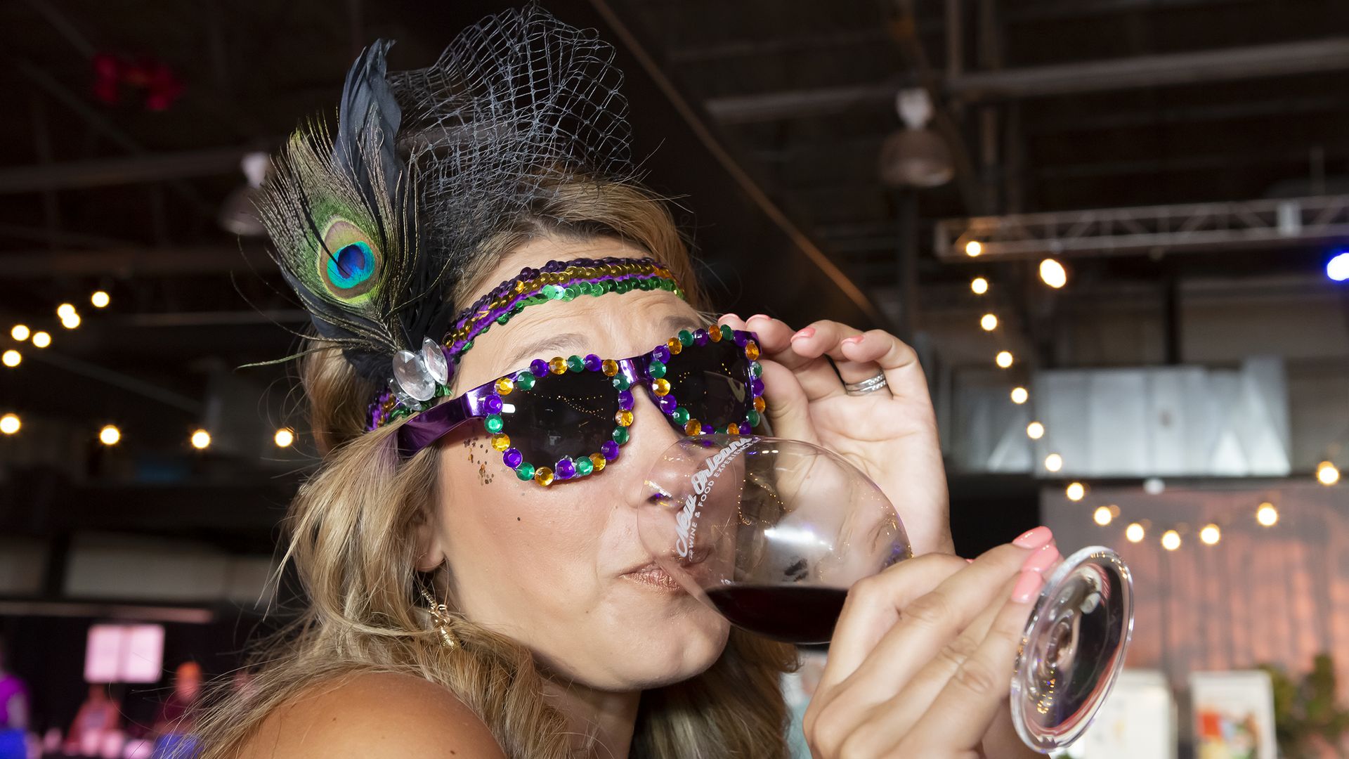 A woman sips a glass of red wine. She's wearing purple sunglasses bedazzled with purple, good and green jewels, and wearing a sequin headband with an ostrich feather.