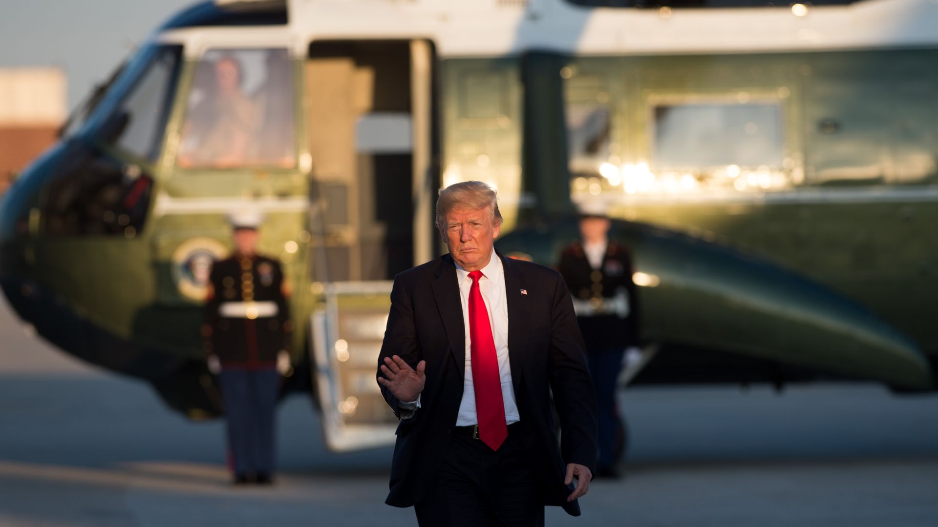 President Trump walking away from a helicopter with his right hand raised, wearing a red tie and a dark suit