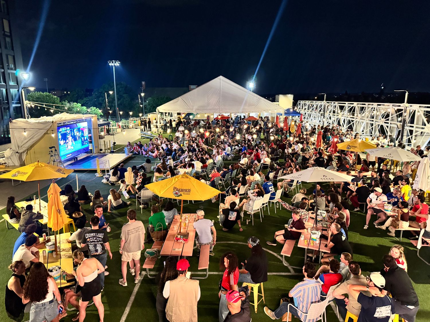 Nighttime outdoor event with a large crowd sitting on grass and picnic benches, watching a big screen on the left. A white canopy tent, string lights, and yellow umbrellas decorate the area.