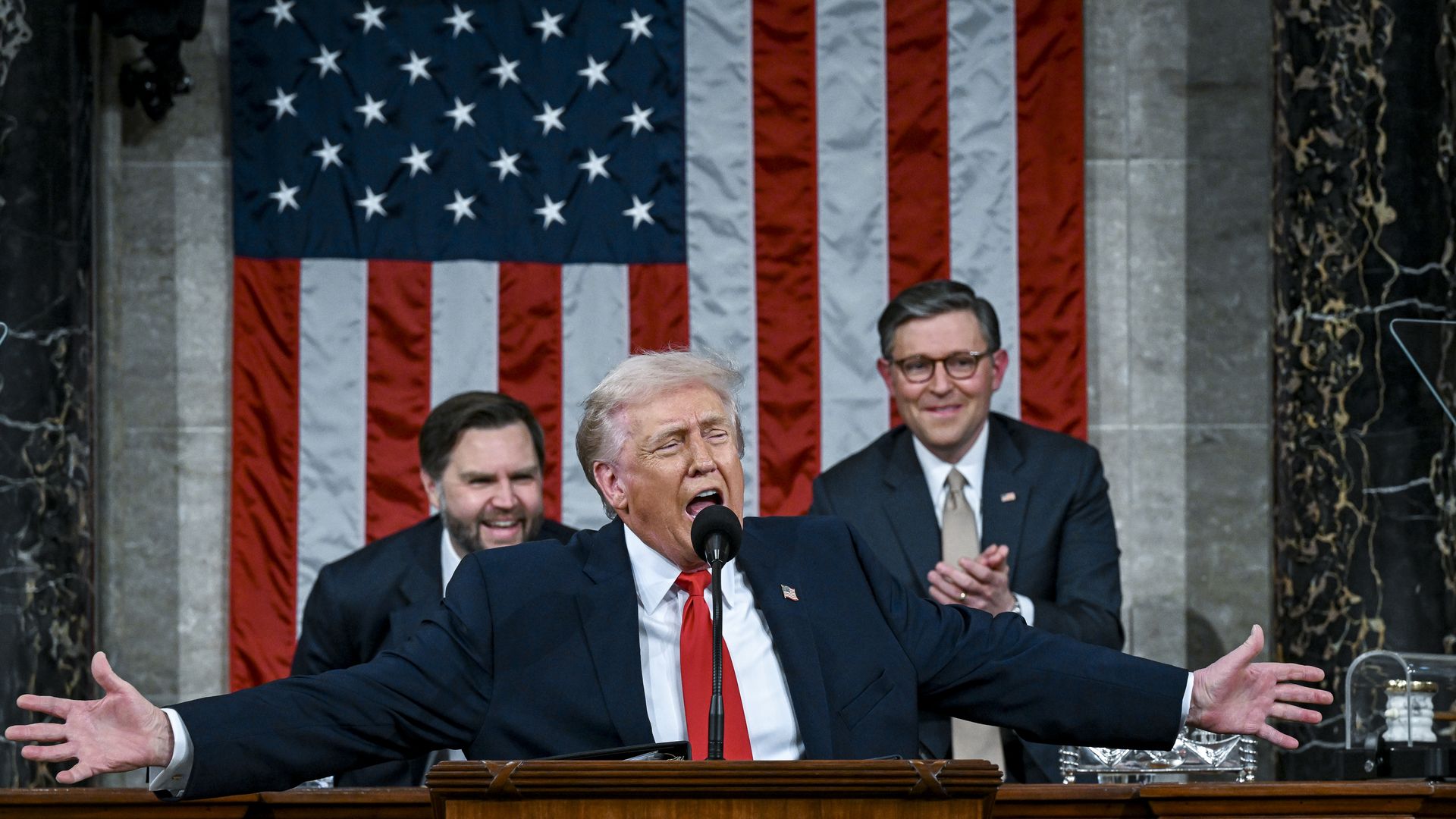 Trump speaking into a microphone with his arms spread wide wearing a navy suit jacket with an American flag pin, white collared shirt and red tie while Speaker Mike Johnson and Vice President JD Vance, standing behind the president, clap and smile