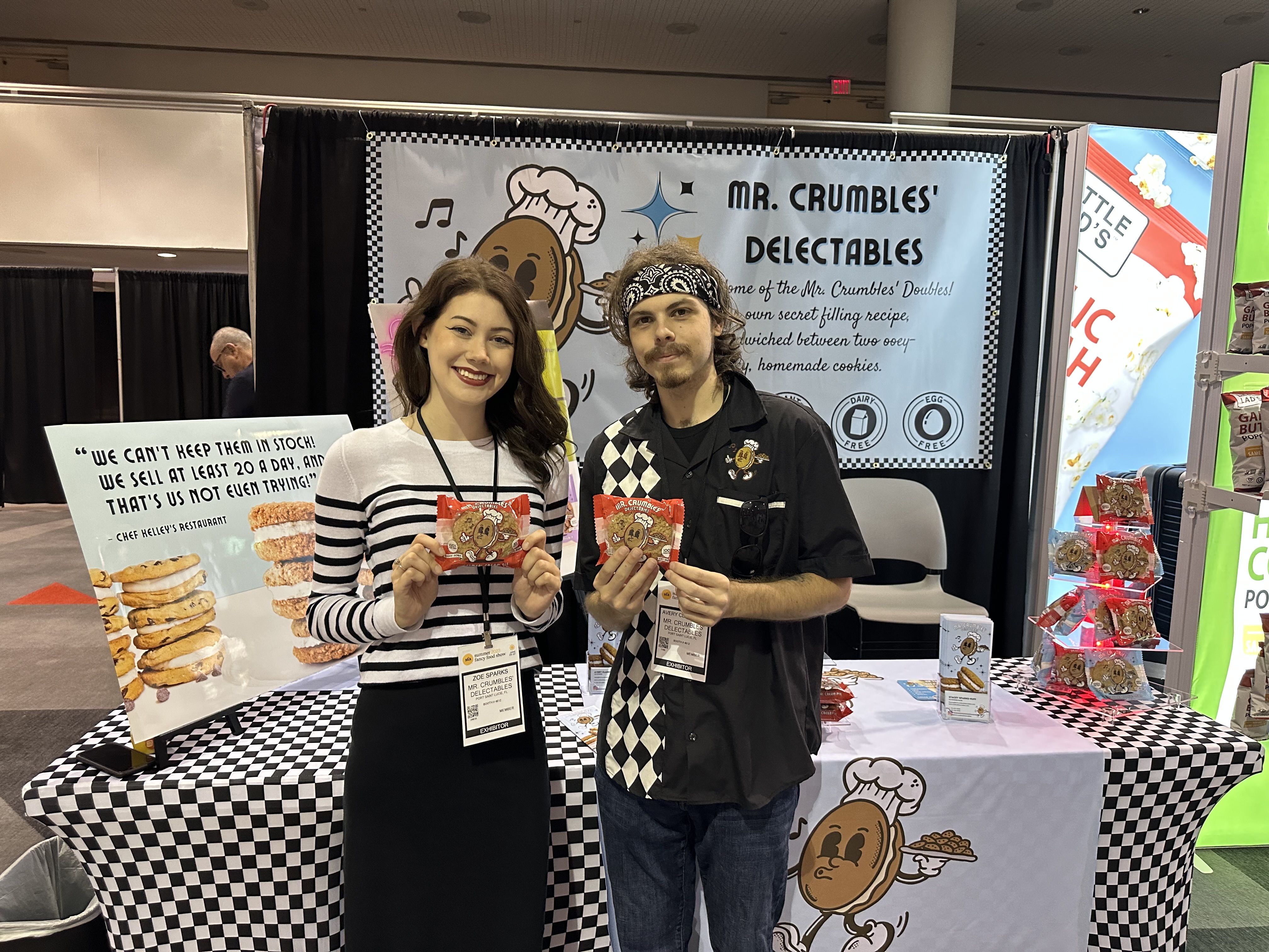Two representatives of Mr. Crumbles' Delectables pose with their cookies in front of a trade show booth.