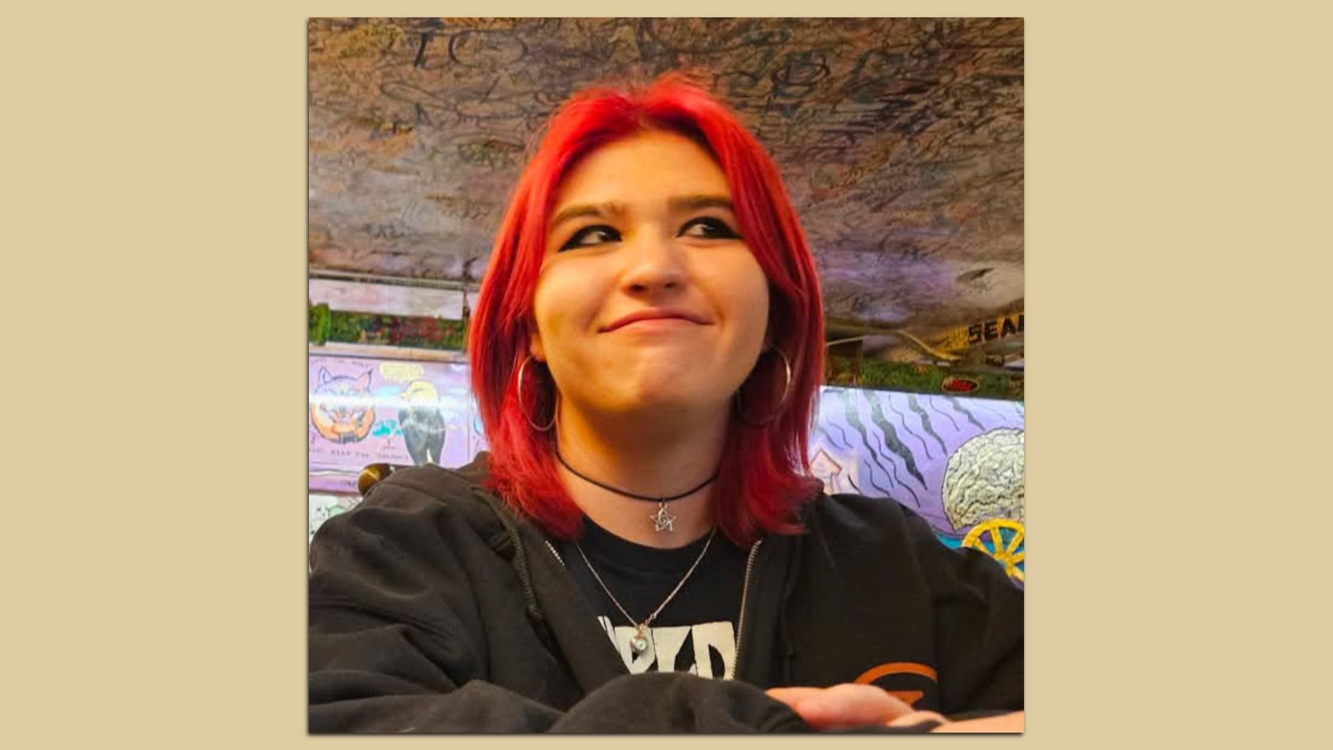 Young person with bright red hair smiles slightly, wearing hoop earrings, black choker with star pendant, black hoodie, and a graphic tee, indoors with colorful graffiti walls.