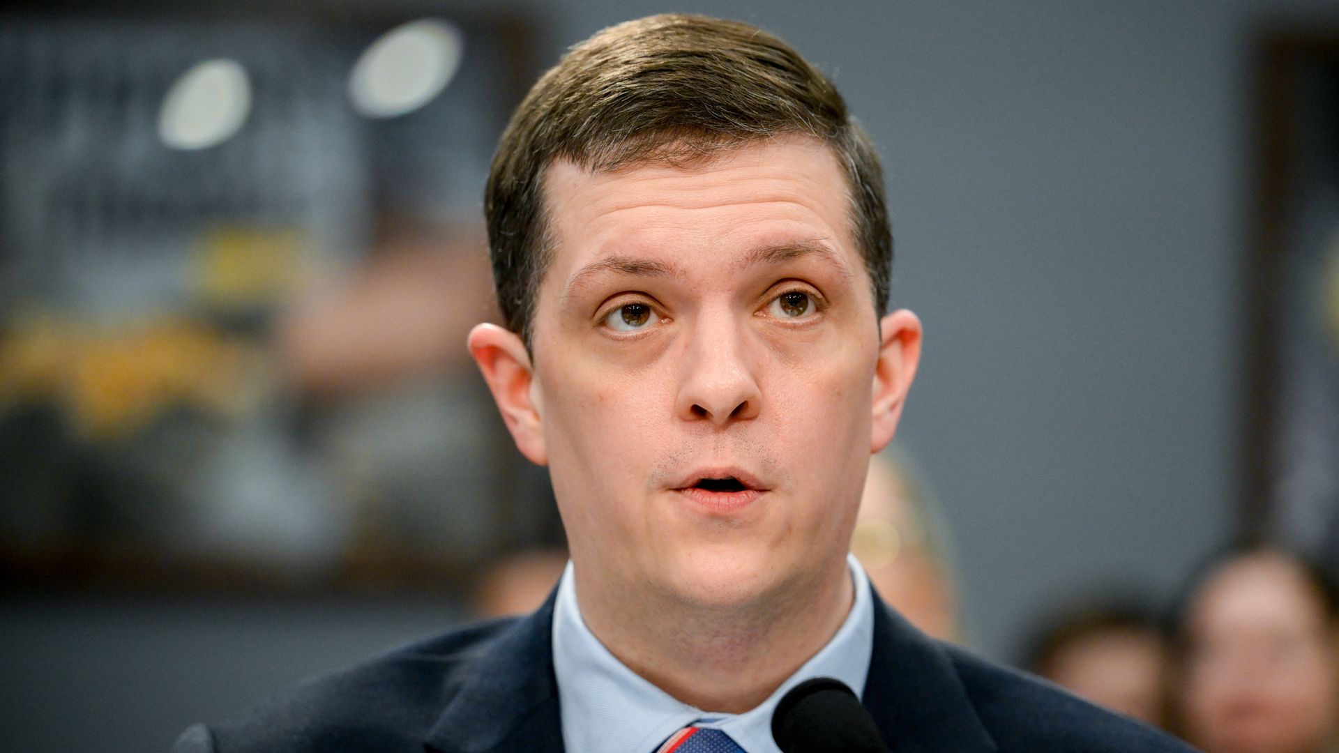 A man wearing a suit talking into a microphone during a congressional hearing. 