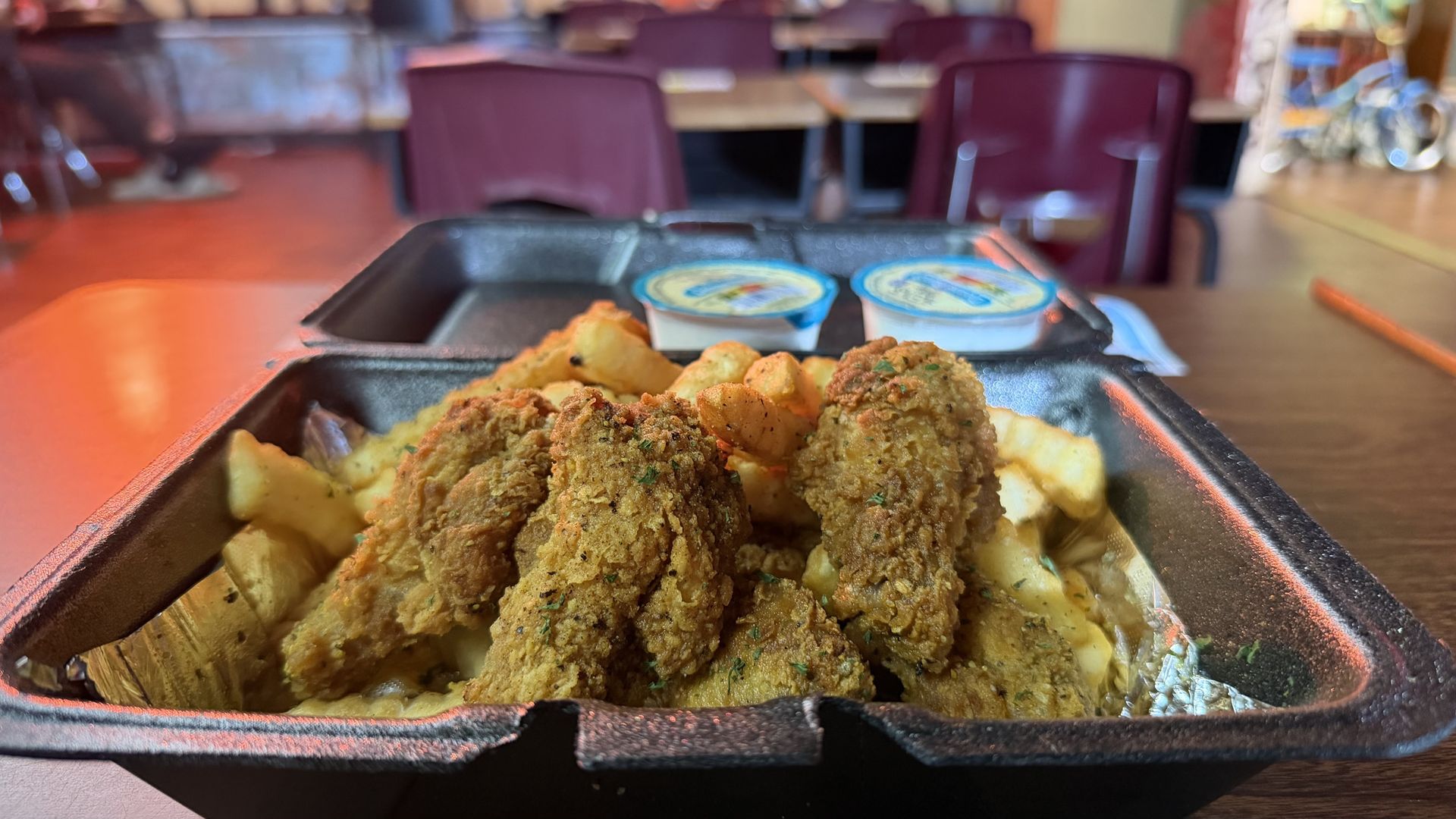 Takeout tray with crispy fried chicken and fries, two sauce cups in the background, set on a table in a casual restaurant with purple chairs.