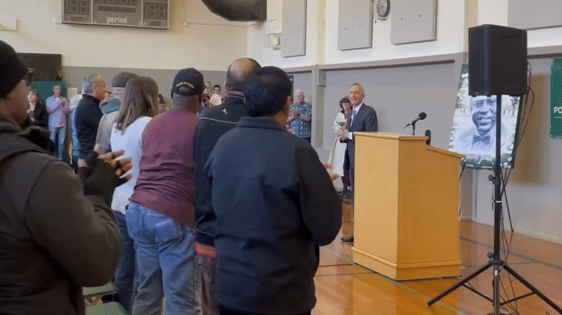 A gif of a person standing behind a lectern and then approaching another person to shake their hand.