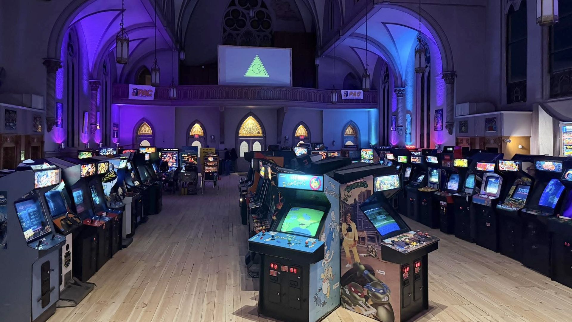 Arcade room inside a cathedral with rows of vintage game machines, purple and blue uplighting on arches, a wooden floor, and stained glass windows at the far end.