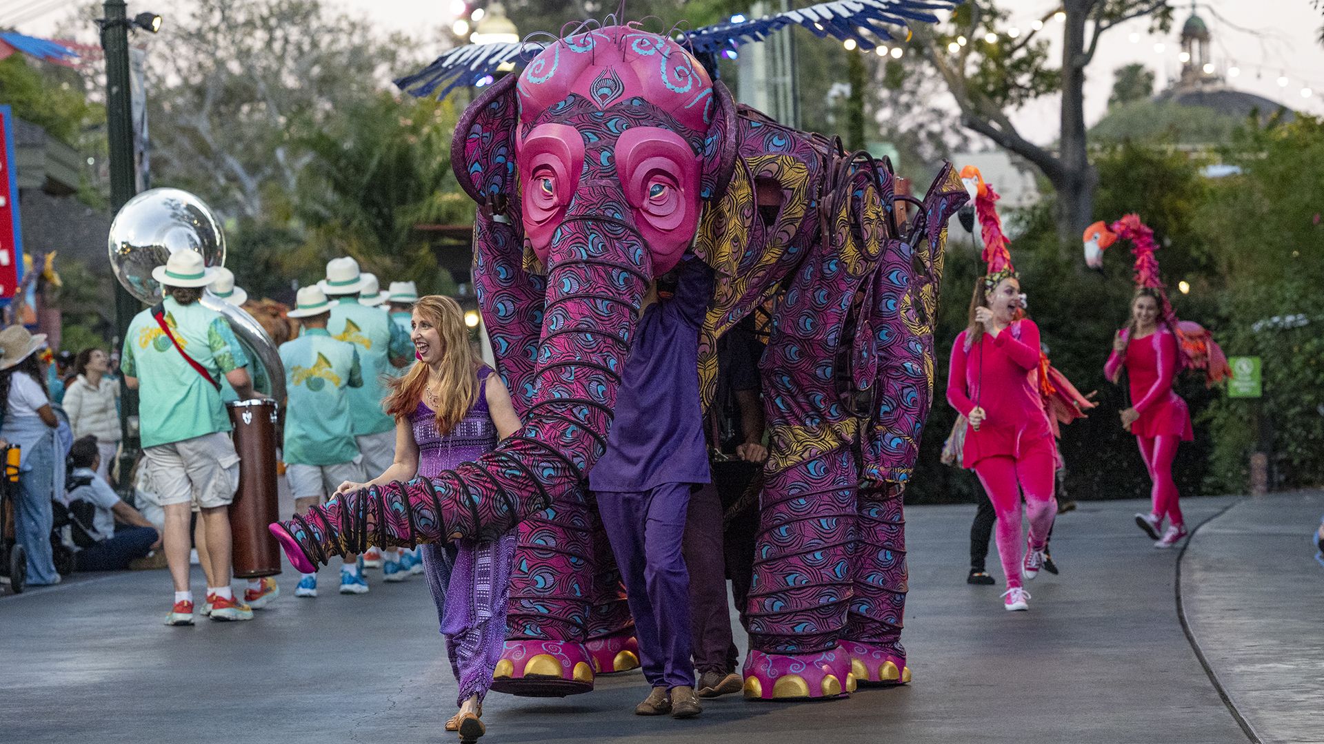 Colorful street parade with a large pink elephant costume center, dancers in bright outfits, musicians in teal shirts, and onlookers along the road under string lights.