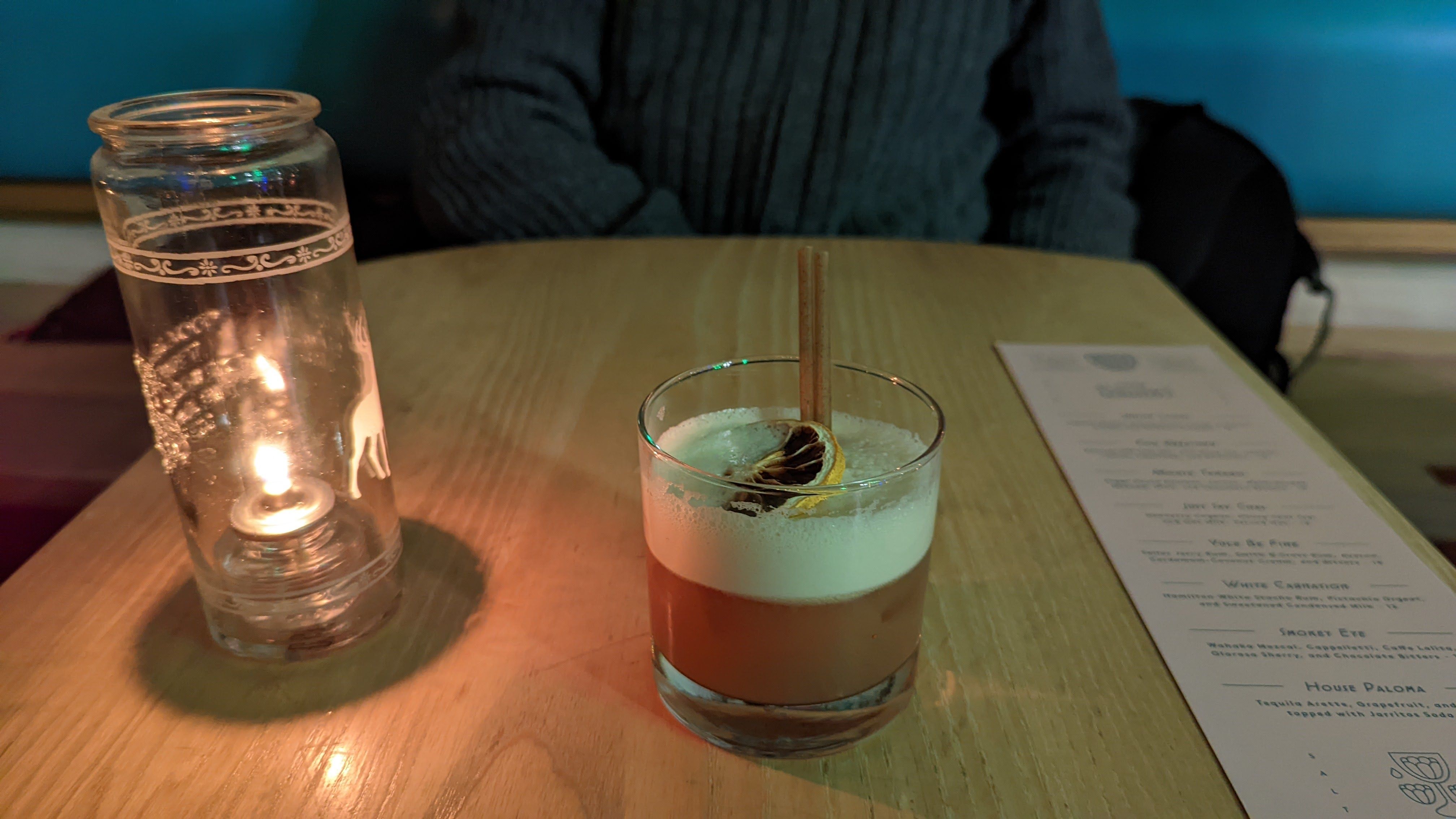 A cream-topped cocktail sits on a table next to an oil lamp.