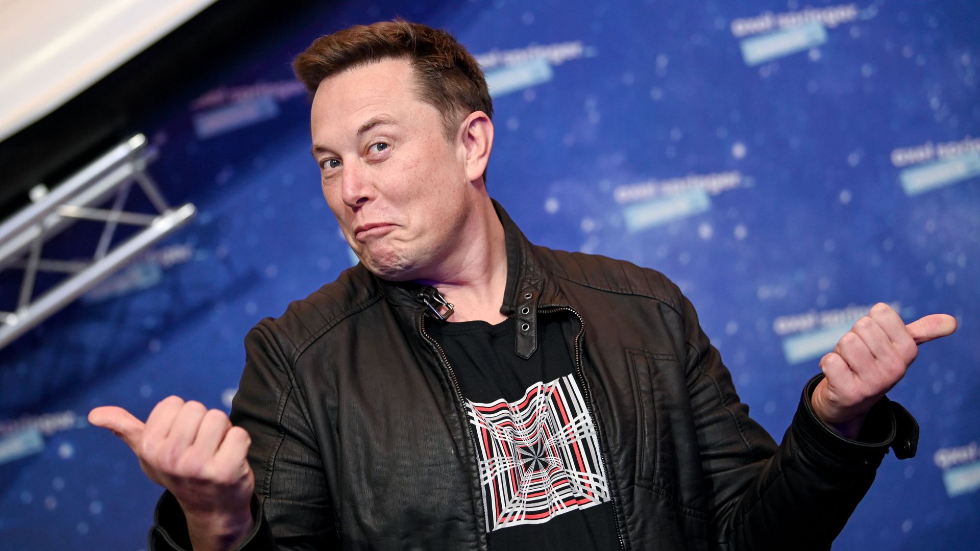 A man in a black leather jacket makes a funny face with pursed lips, raised eyebrows, and hands gesturing, against a blue, starry backdrop.